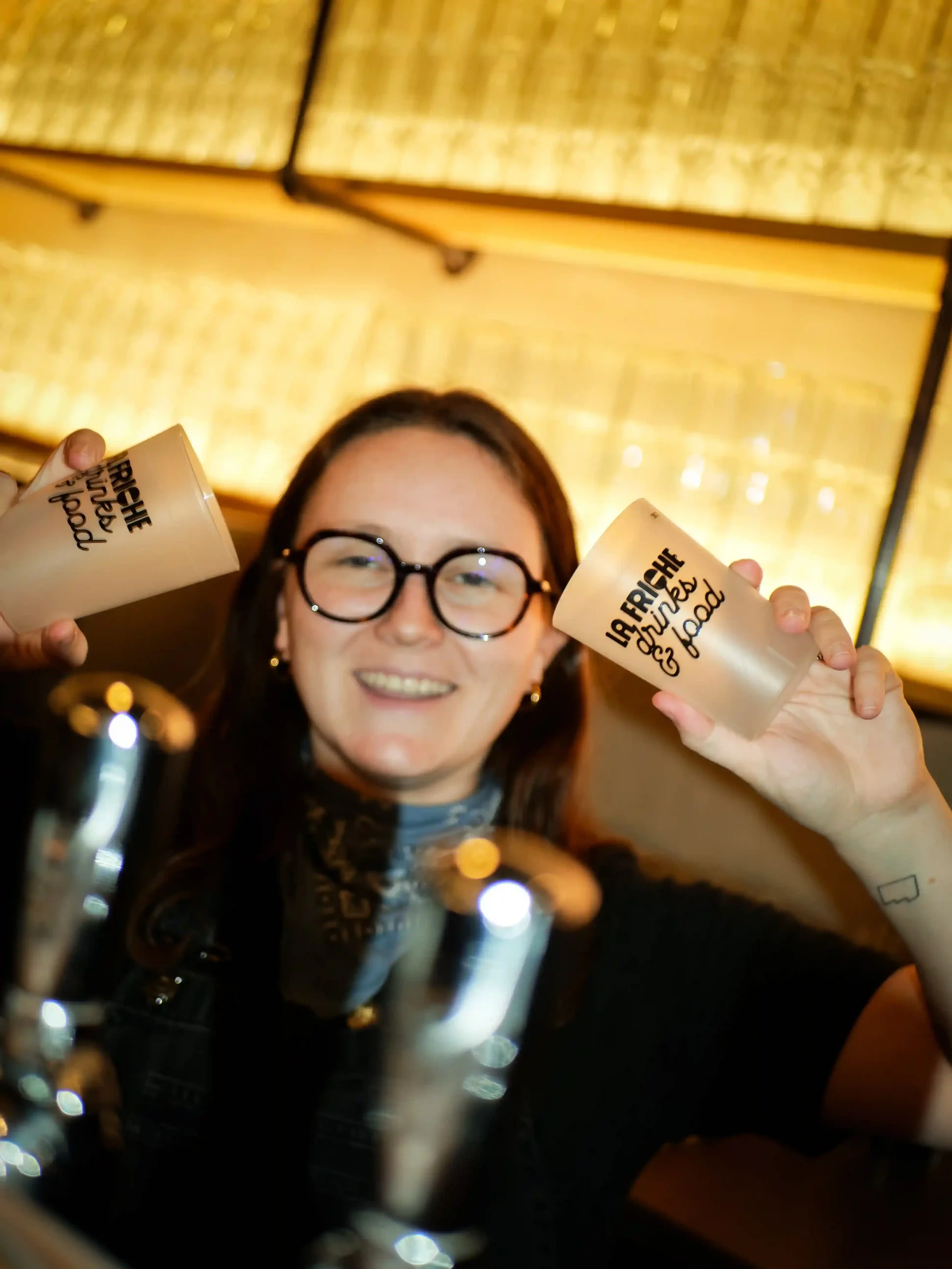 Une femme souriante avec des lunettes noires tient deux gobelets en plastique, un dans chaque main, dans une ambiance de bar ou restaurant.