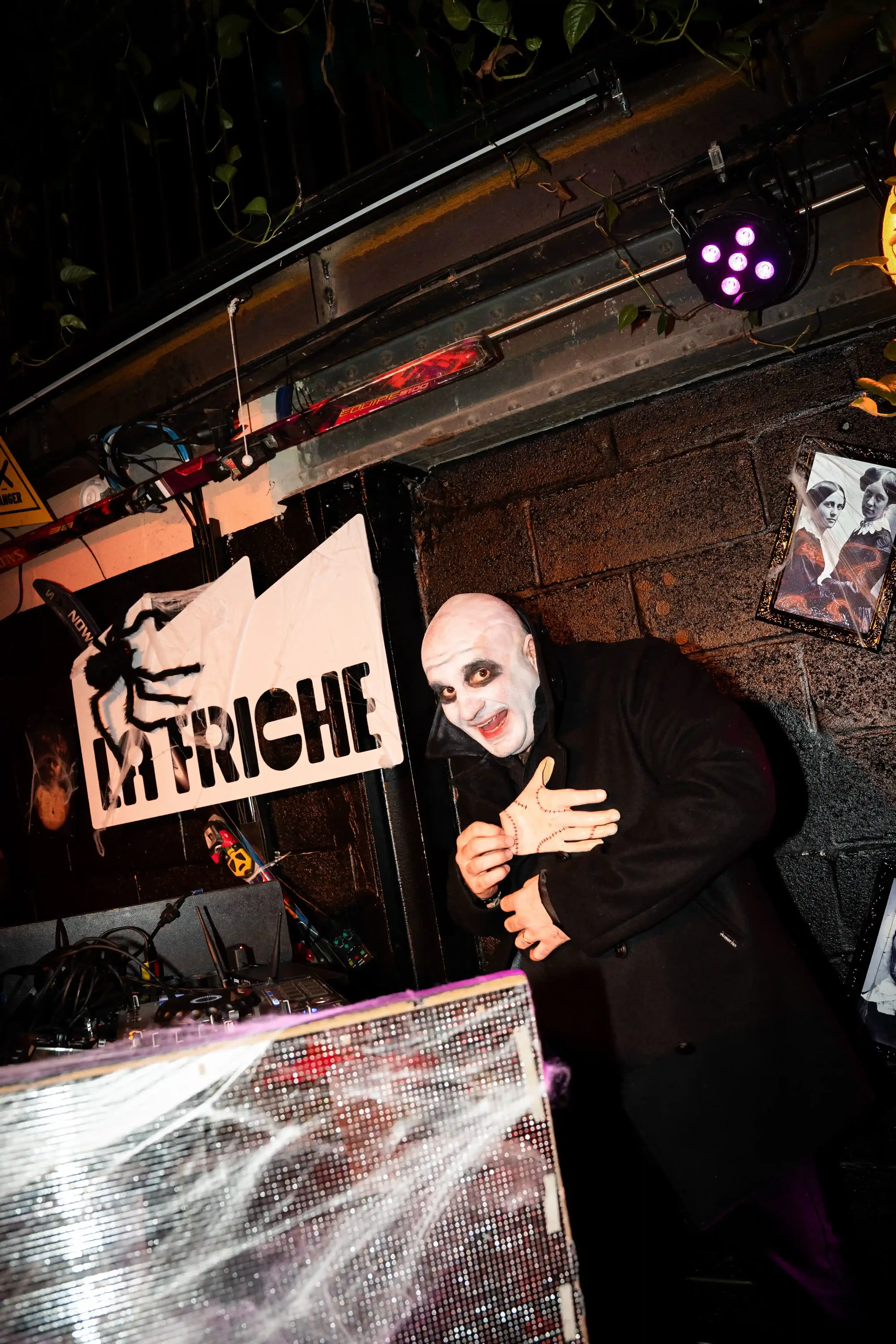 Homme déguisé en Dracula avec un visage blanc, maquillage sombre autour des yeux, souriant et posant dans un décor sombre avec une affiche de la Triche et une araignée en plastique suspendue. Variante de photo de soirée avec éclairage violet.