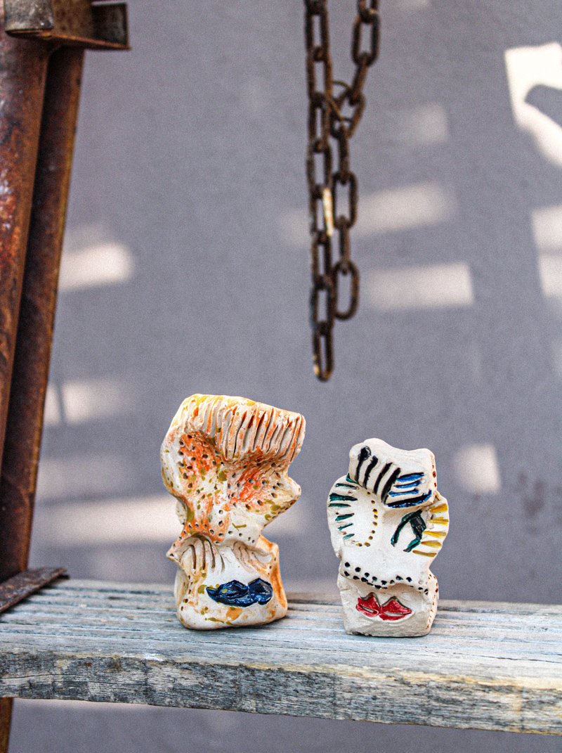 Two small, highly textured ceramic sculptures of abstract faces with colorful glazes by Dimitra Kalantzi, resting on a weathered wooden plank at Plastico Gallery.
