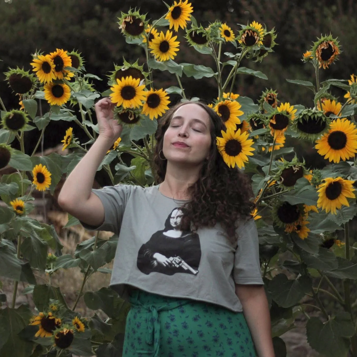 Portrait of artist Agni Roussou standing among tall blooming sunflowers with her eyes closed, a featured creator at Plastico Gallery.