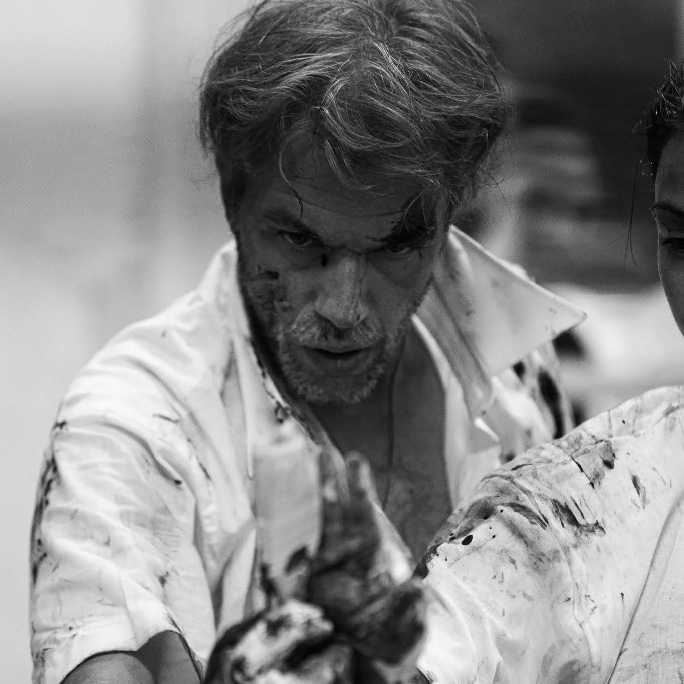 Black and white action portrait of artist Vasillis Kavouridis with a focused expression, his face and white shirt covered in dark paint splatters, performing at Plastico Gallery.