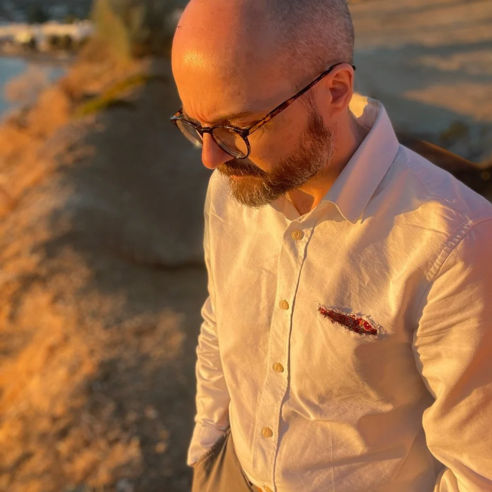 Portrait of Spiros Baras looking thoughtfully downward in warm golden hour sunlight, wearing a white shirt and glasses, a featured artist at Plastico Gallery.