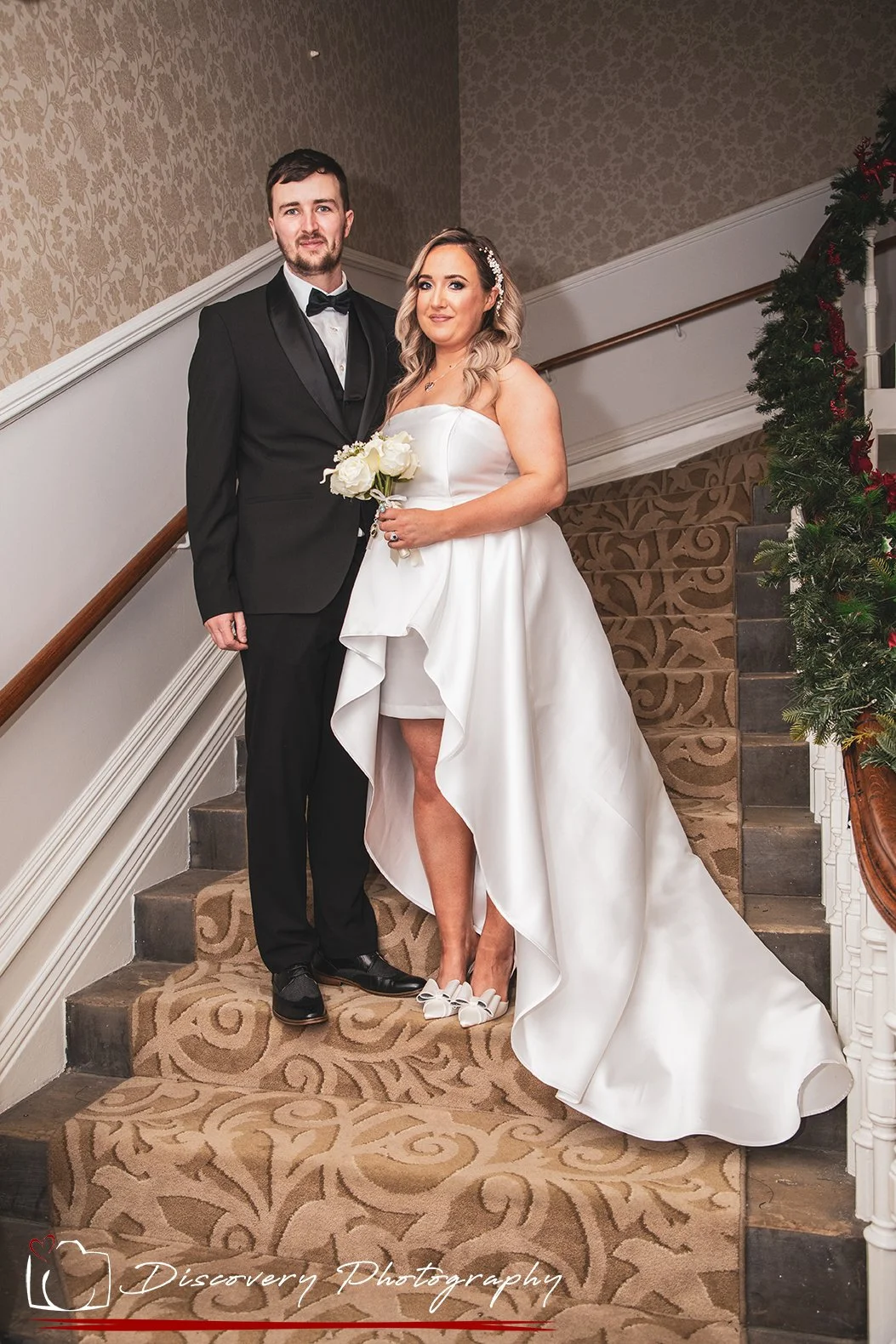 A bride wearing a strapless white wedding gown and holding a bouquet of white roses, standing on a staircase with a groom dressed in a black tuxedo and bow tie, at a wedding celebration decorated with greenery and red ribbons.