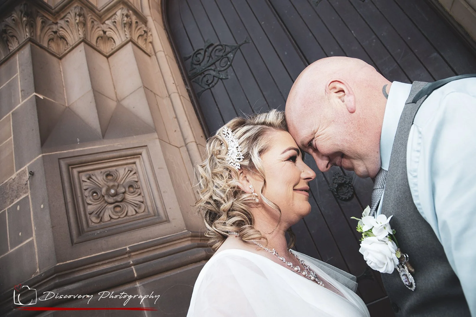 A bride and groom touching foreheads and smiling at each other outside a building with ornate stonework and a dark wooden door.