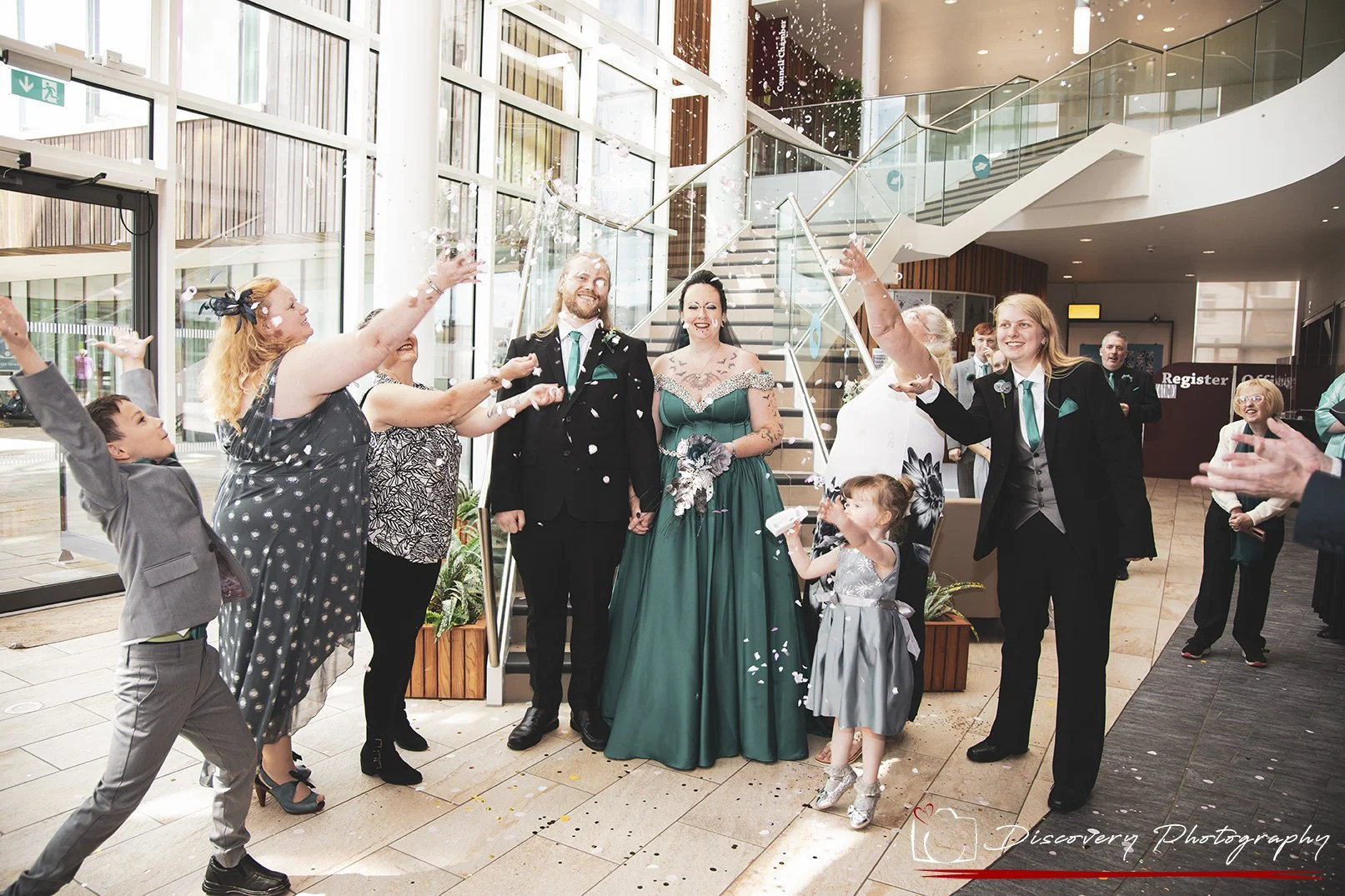 A wedding celebration with a group of people cheering and throwing confetti inside a building with glass walls and a staircase.