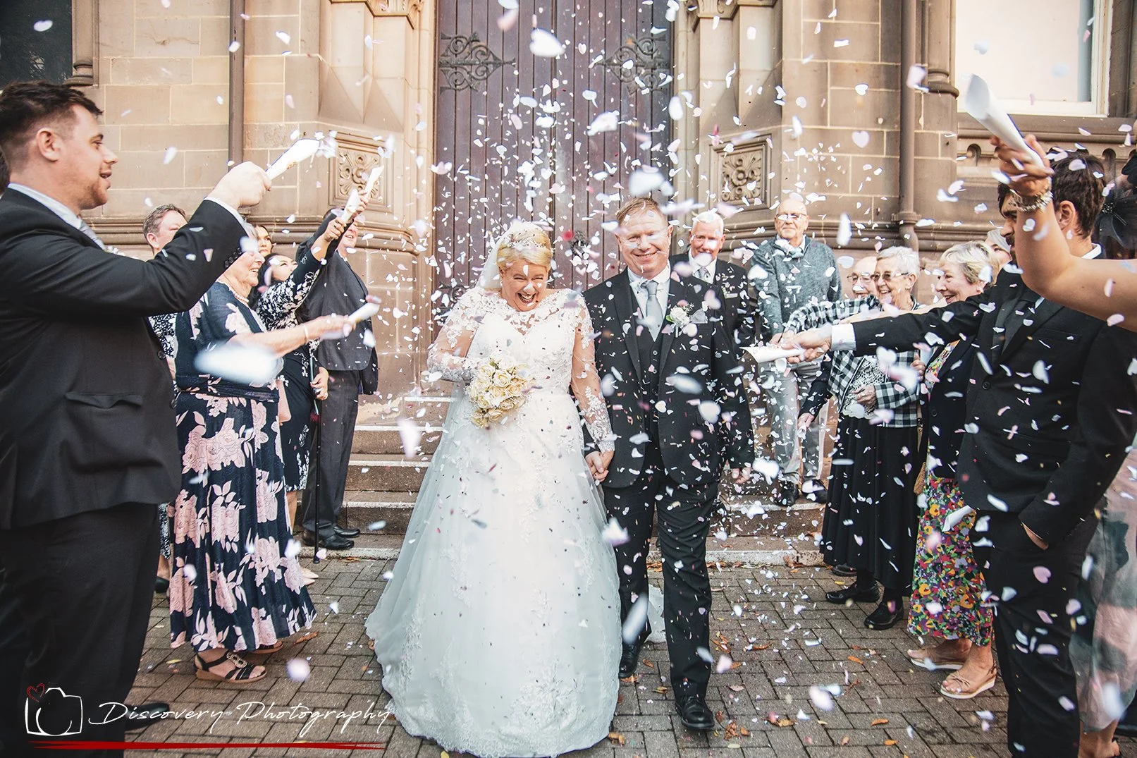 A bride and groom walking out of a church, being showered with confetti by friends and family on their wedding day.