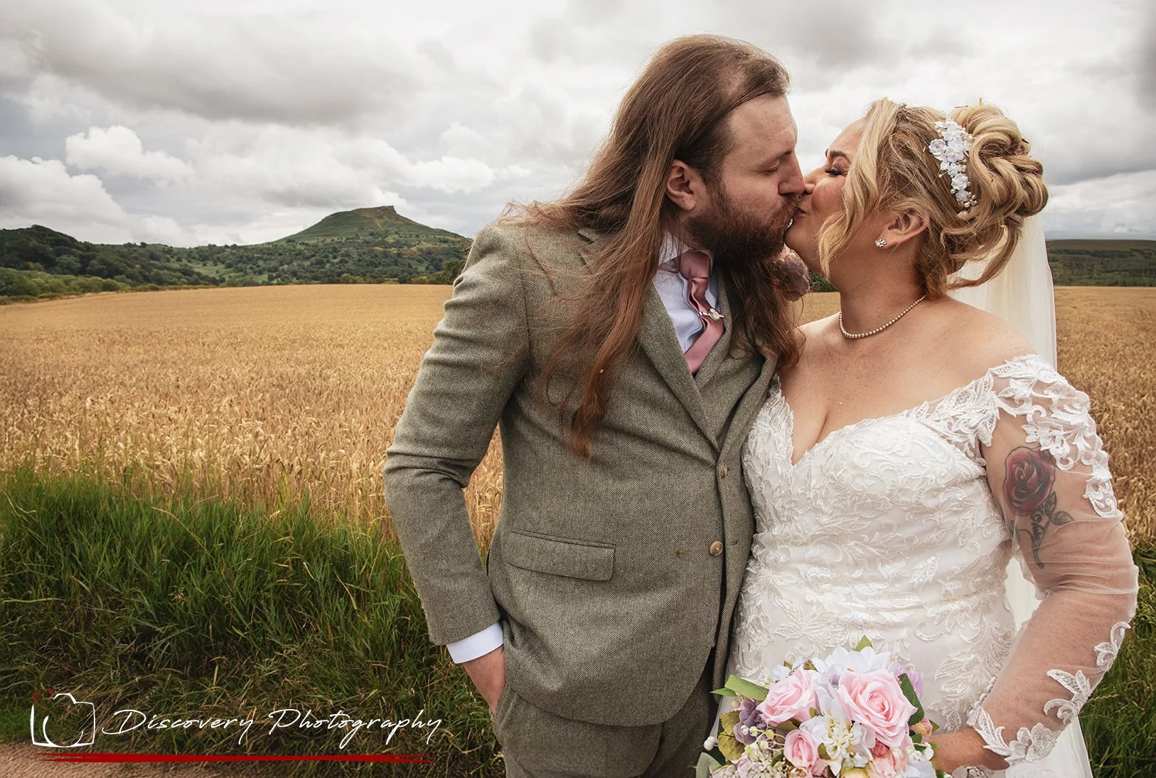 A newlywed couple with long hair sharing a kiss in a field with a mountain in the background, bride holding a pink and white bouquet.