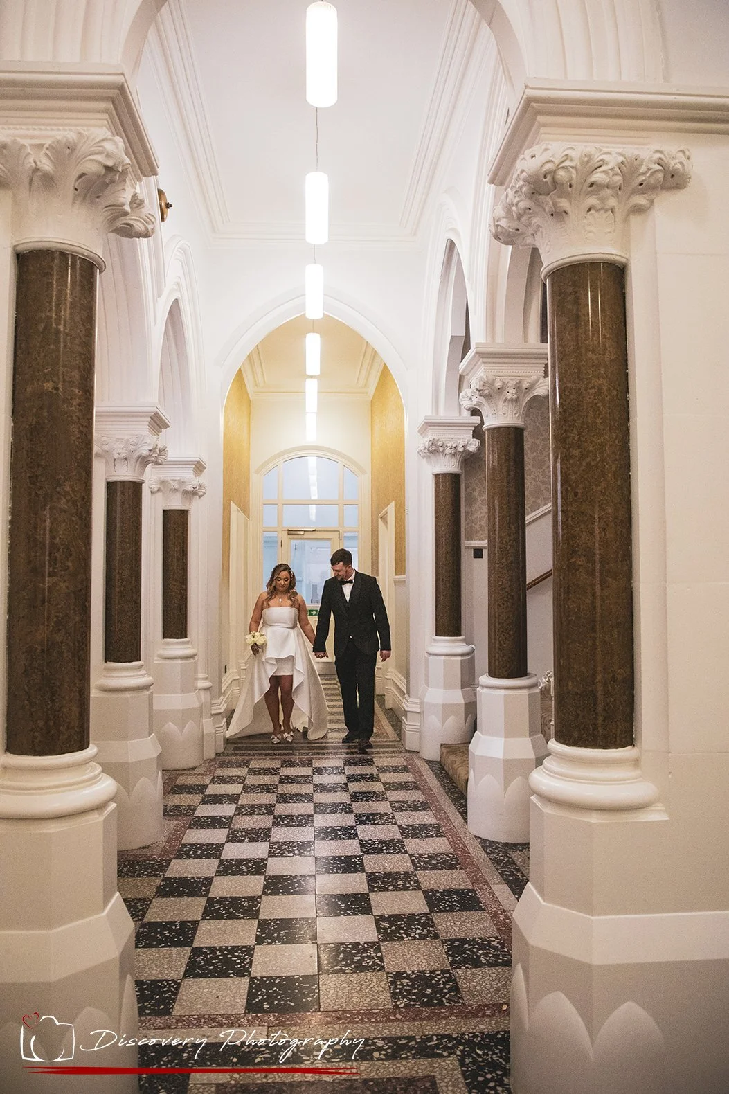 Bride and groom walking hand in hand through the elegant hallway at Middlesbrough Registry Office, framed by grand columns and a patterned tiled floor, captured by a North East wedding photographer