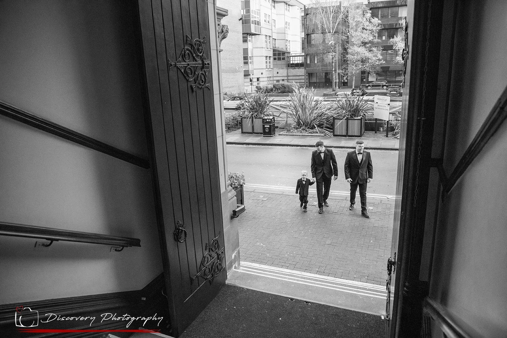 Black and white photograph of the groom arriving at Middlesbrough Registry Office with the couple’s children, captured from inside the doorway by a documentary-style North East wedding photographer