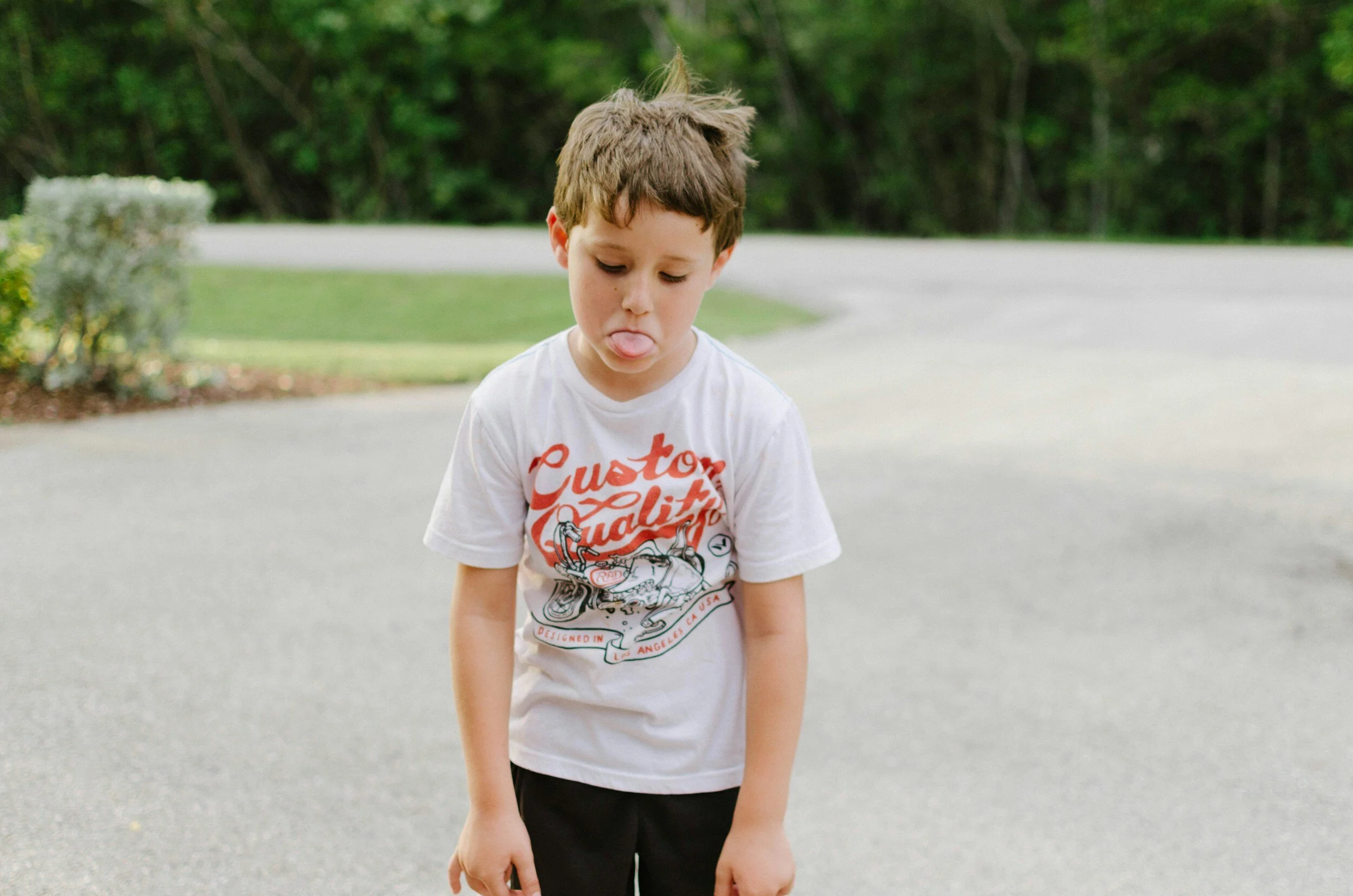 Young boy standing outdoors on a paved surface with trees and bushes in the background, sticking out his tongue.