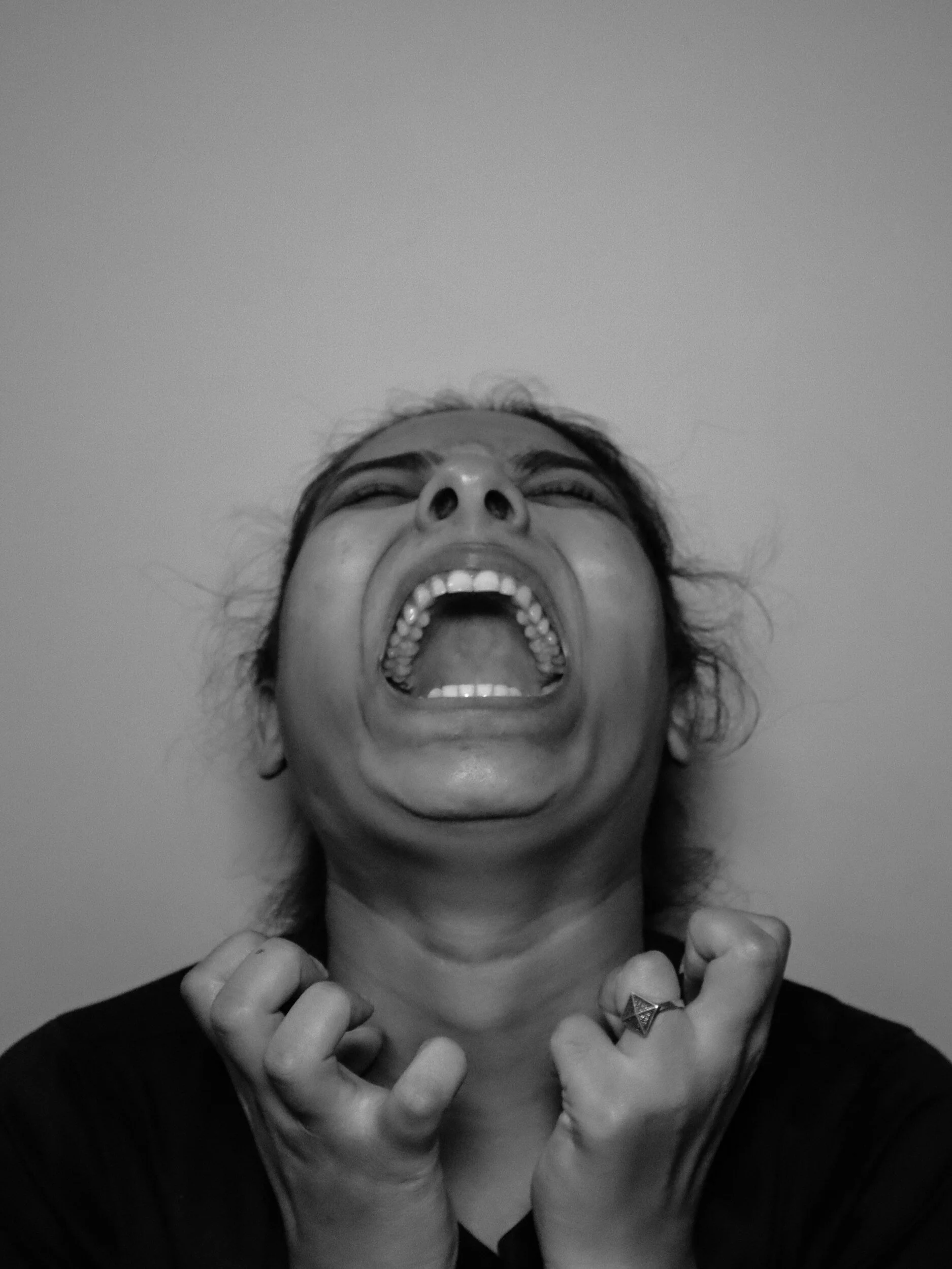 Black and white photo of a woman with short hair, expression of distress or pain, mouth wide open, hands clenched near her neck, wearing a ring on her left hand.