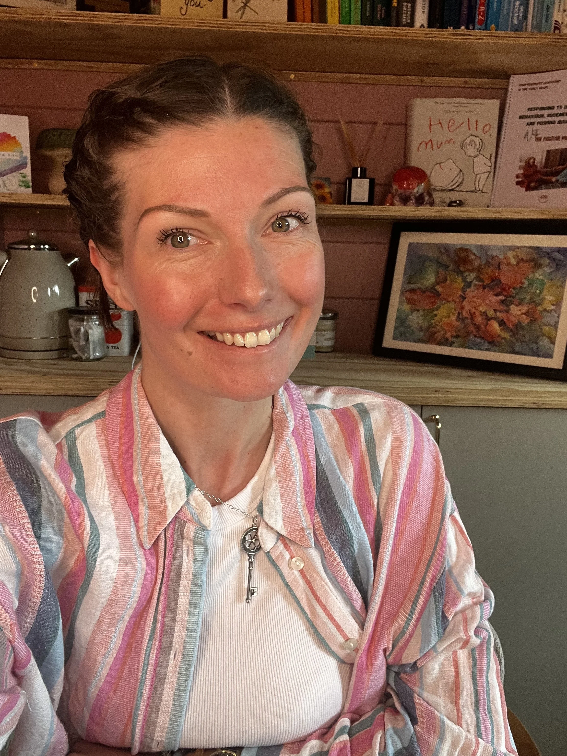 Close-up of a smiling woman with short brown hair, wearing a multicolored striped shirt and a necklace with a key pendant, sitting in front of a wooden shelf with books, framed art, and decorative items.