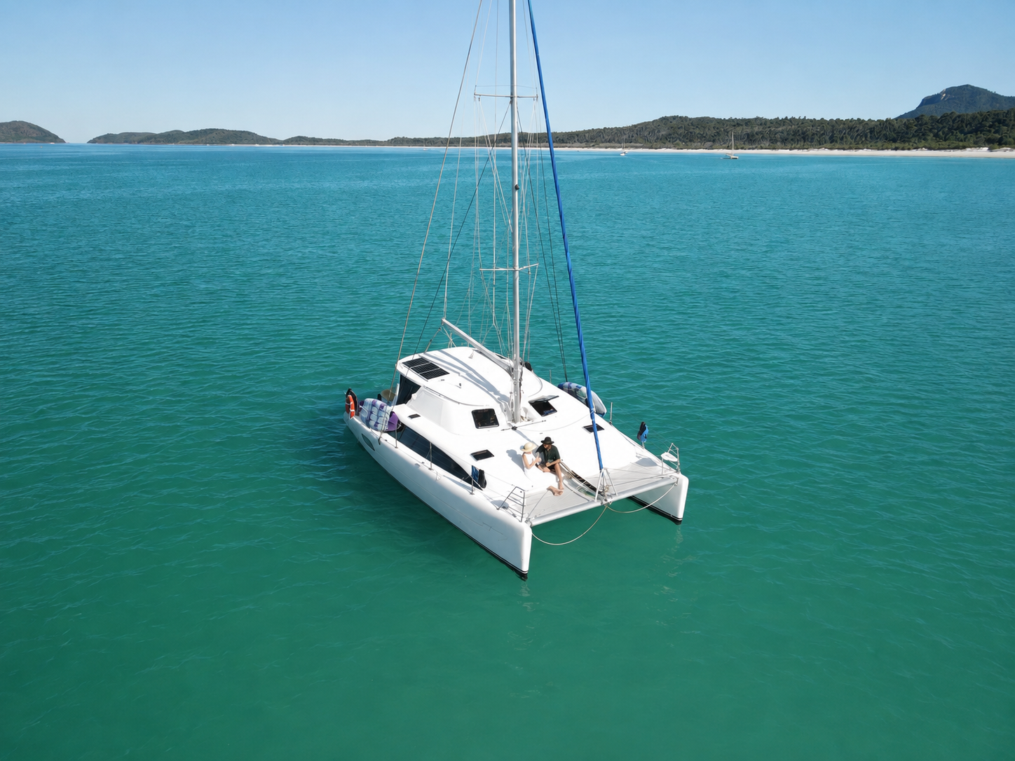 A white sailing catamaran with two people sitting on the deck, floating on calm turquoise water near a distant shoreline with green hills and islands under a clear blue sky.
