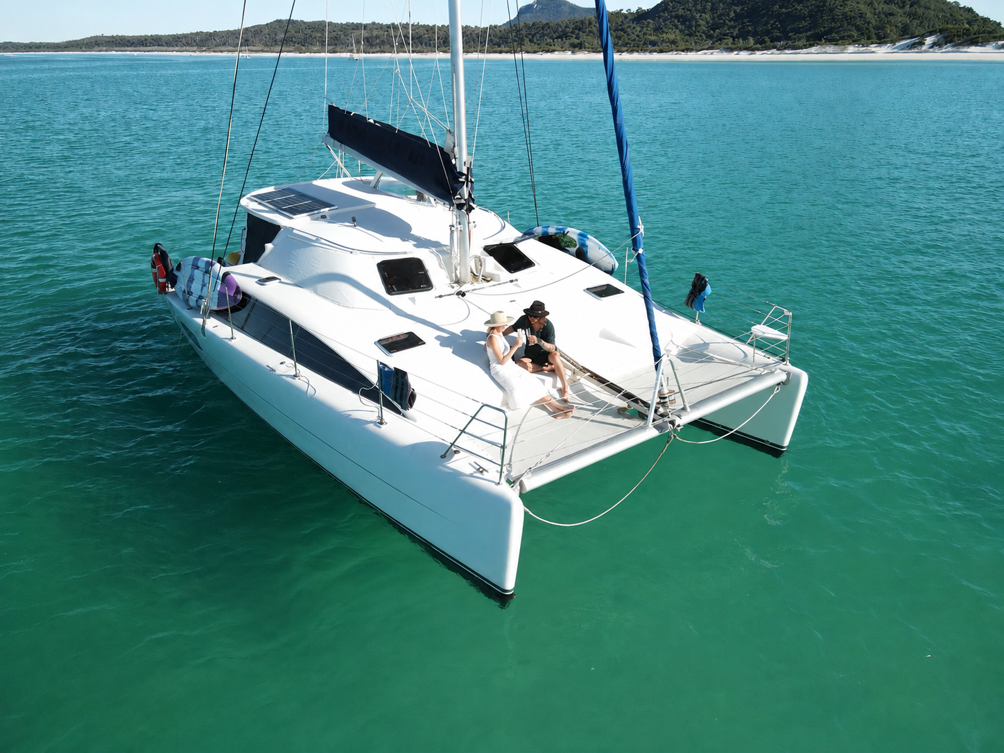 A white sailing yacht floating on clear turquoise water with two people sitting on the deck, enjoying a conversation, against a background of a distant shoreline with trees and hills.
