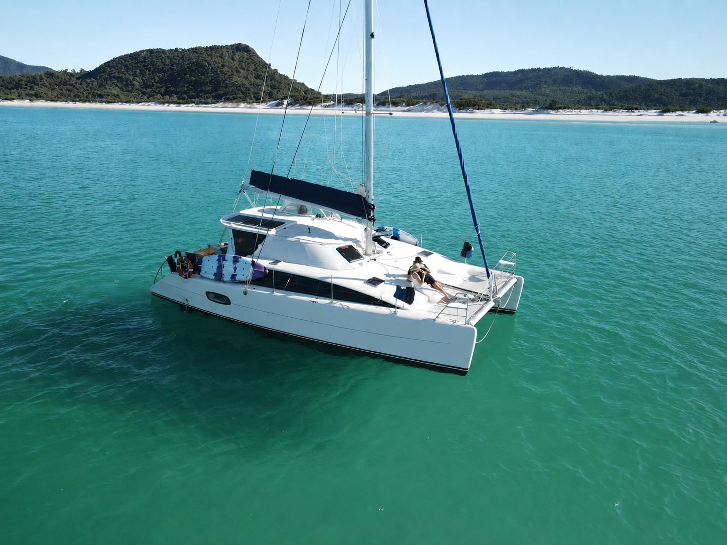 A white sailboat on clear turquoise water with two people relaxing on the deck, in front of a green, hilly landscape under a blue sky.