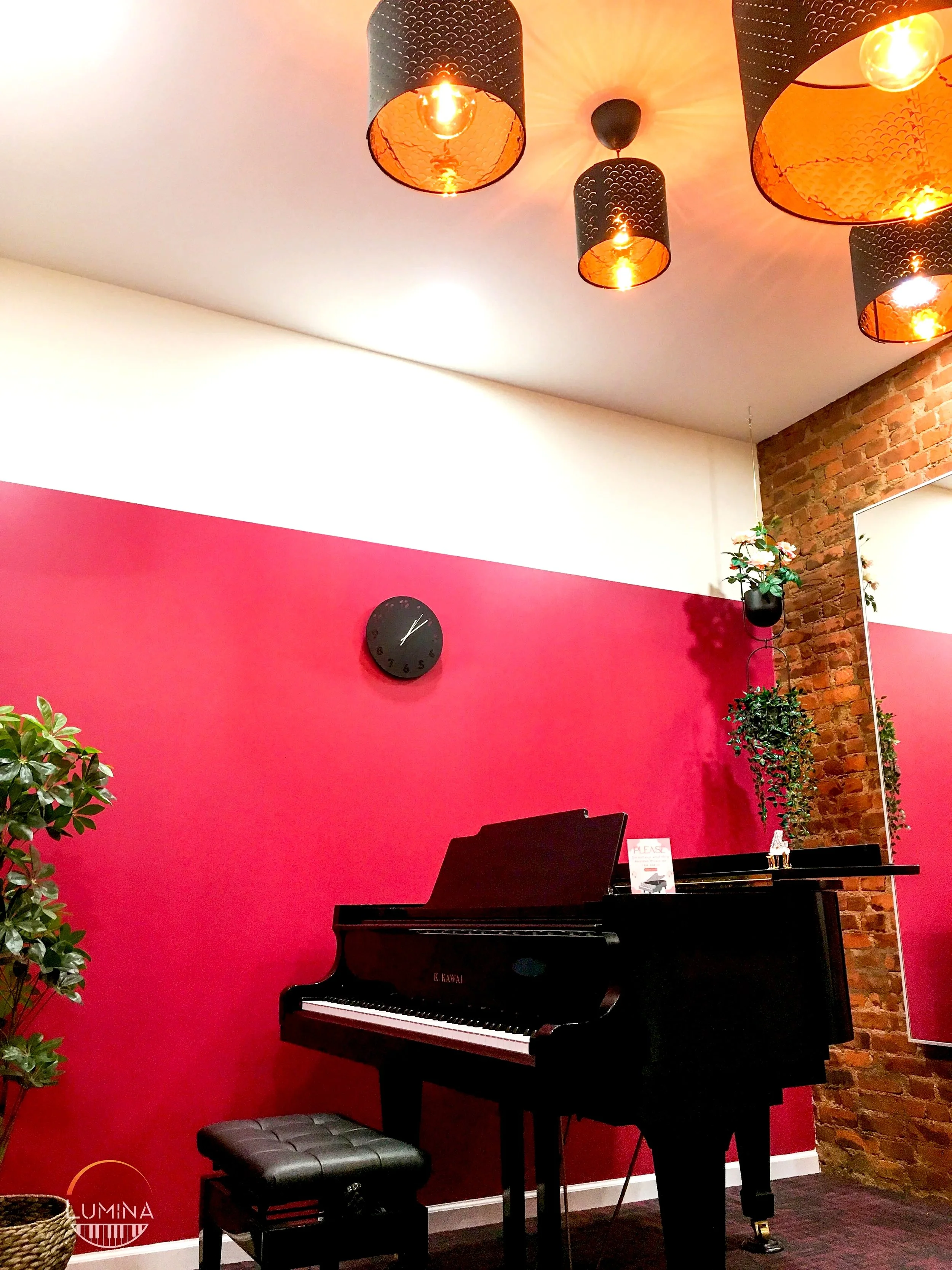 A Kawai black grand piano is situated in a room with a pink wall, a white ceiling, and an exposed brick wall. The piano has a small sign and a decorative horse figurine on top. A black piano bench is in front of the instrument, and a plant is visible on the left side. The ceiling features five black pendant lights with orange interiors, hanging above the piano. There is a black clock on the pink wall and a tall potted plant on the right side.