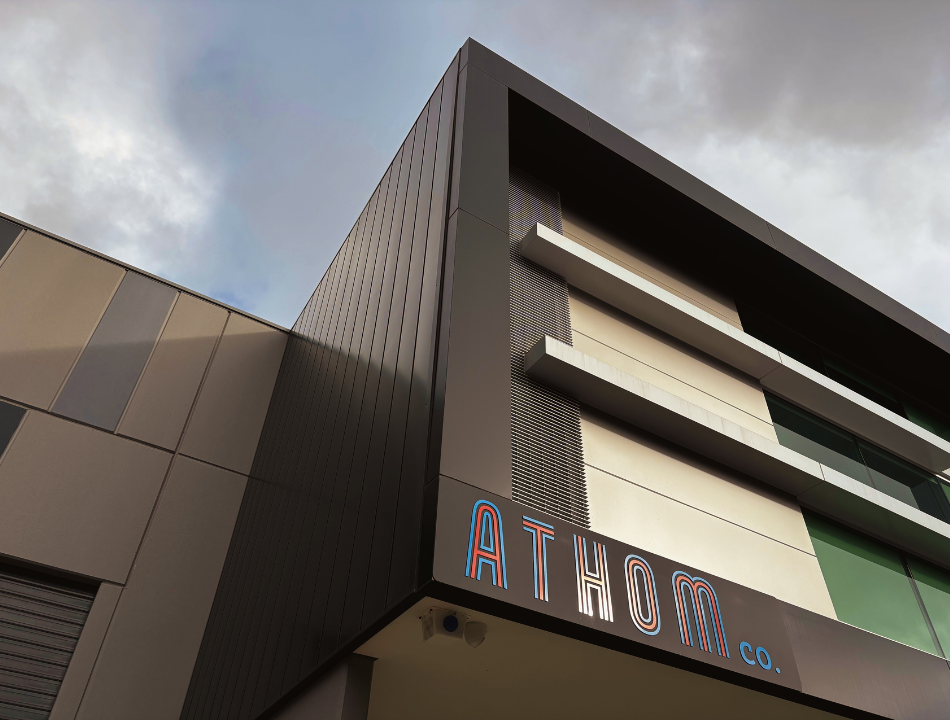 Close-up view of a modern building with a sign reading 'ATHOM CO' at the bottom corner. The building features sleek, dark and beige panels with horizontal metal accents and a cloudy sky in the background.