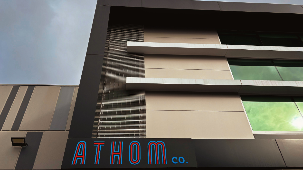 Modern office building with large windows reflecting green sky, with a sign reading 'ATHOM co.' in blue and red letters.