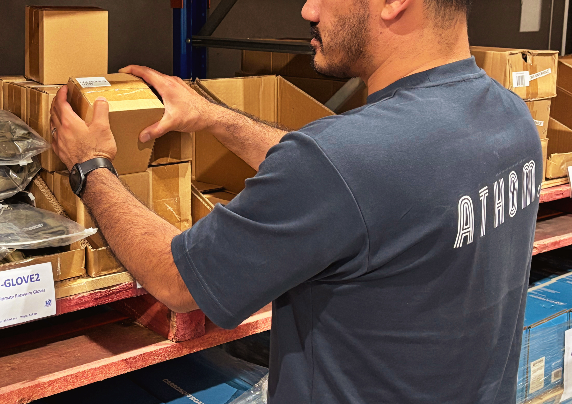 A man wearing a dark blue t-shirt with white text on the back is examining a small cardboard box on a store shelf. The shelf contains various boxes, some labeled with product information.
