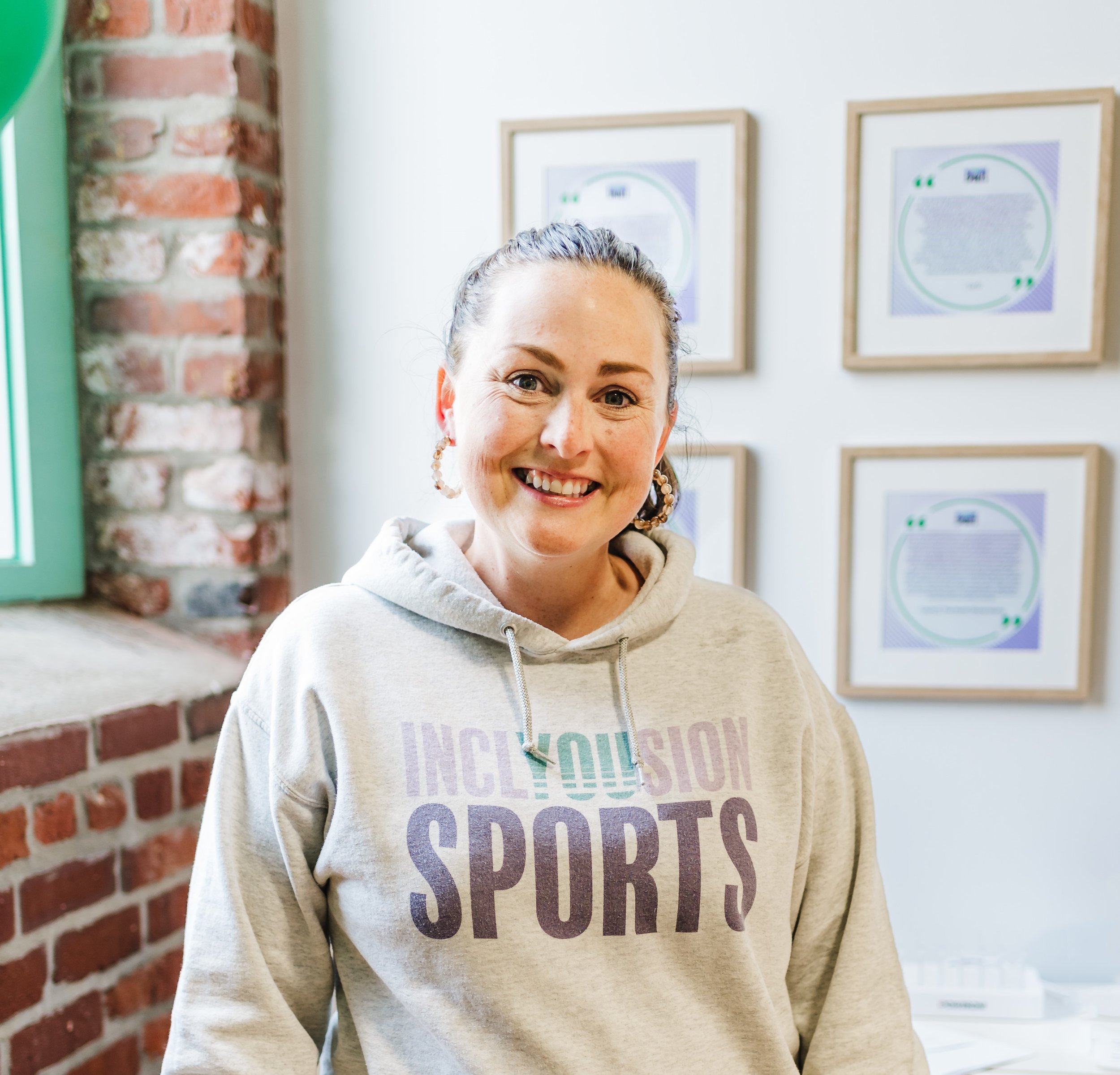 A smiling woman with brown hair tied back, wearing a beige hoodie with purple and green text, standing indoors near a brick wall and framed posters with text.