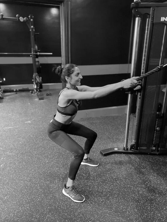 Woman exercising with pulley machine in a gym, performing a squat while holding a cable.