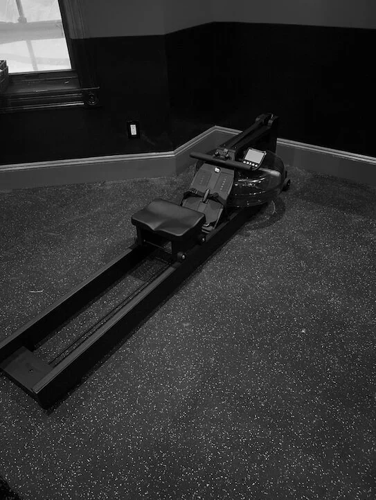 Black and white photo of a rowing machine in a room with dark walls and a textured floor.