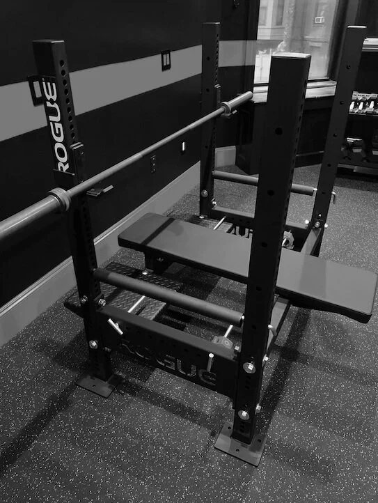 Black and white image of a Rogue bench press with an attached barbell, located in a gym setting.