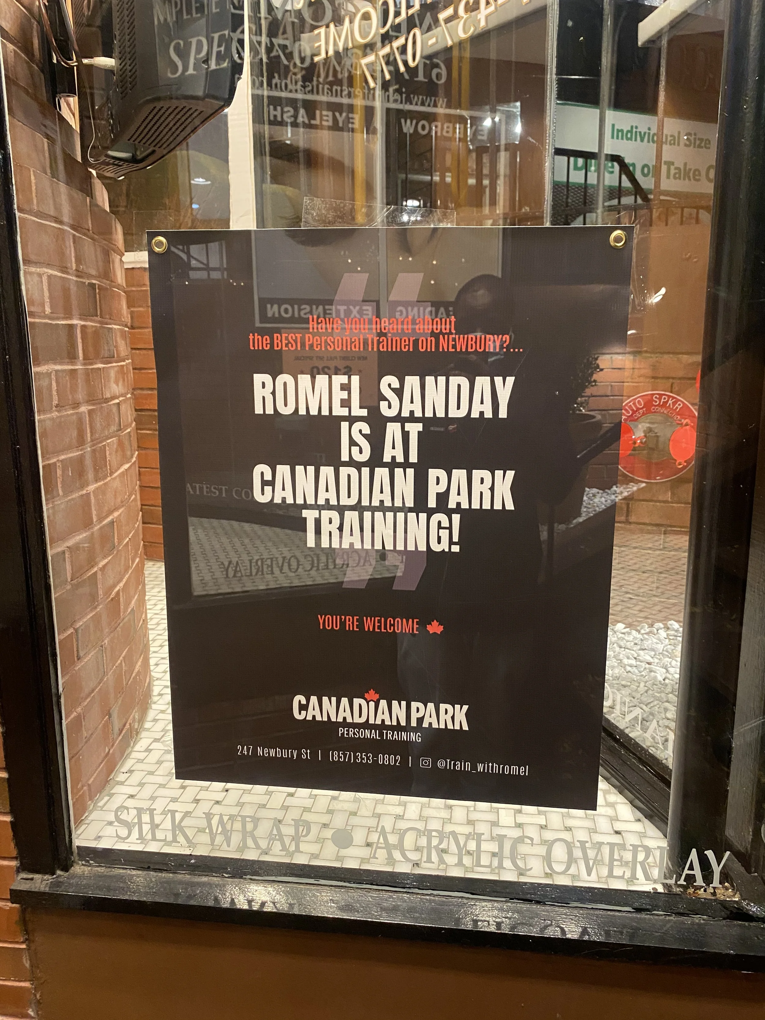 Poster on a window advertising Romel Sanday as a personal trainer at Canadian Park Training, located at 247 Newbury St. Includes contact details and social media handle @train_withromel.