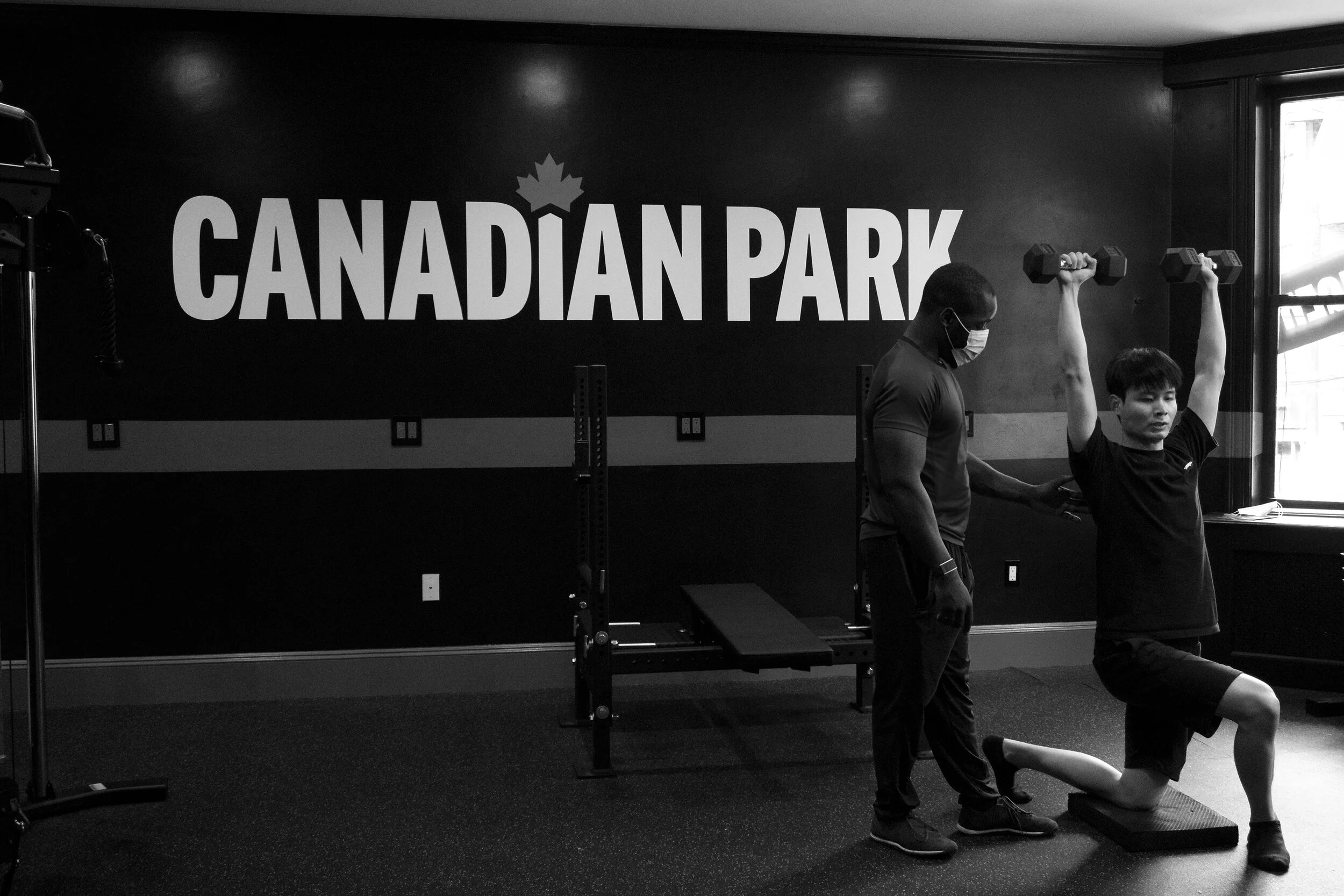A man wearing a face mask assists another man who is doing a lunge exercise with dumbbells at a gym labeled 'Canadian Park.'