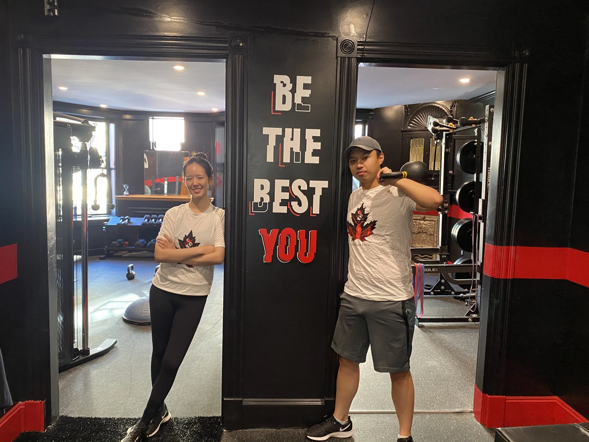Two people standing in a gym, one holding a kettlebell, near a wall with the slogan "Be The Best You."