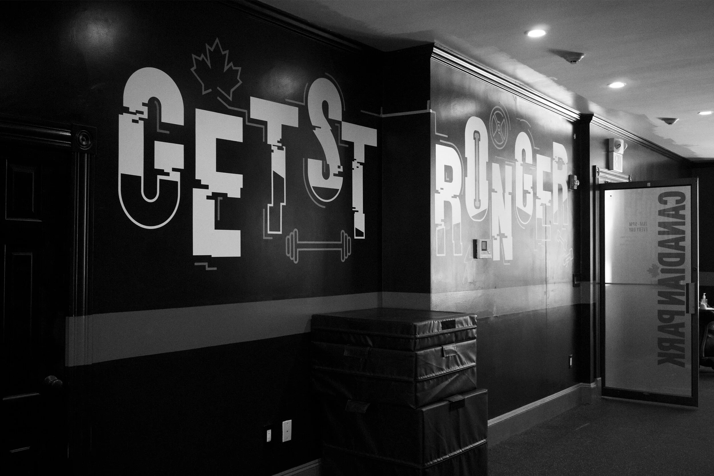 Black and white gym interior with motivational text "Get Stronger" on the wall, featuring a barbell graphic and maple leaf symbol. Stacked gym mats and a door with "Canadian Park" visible.