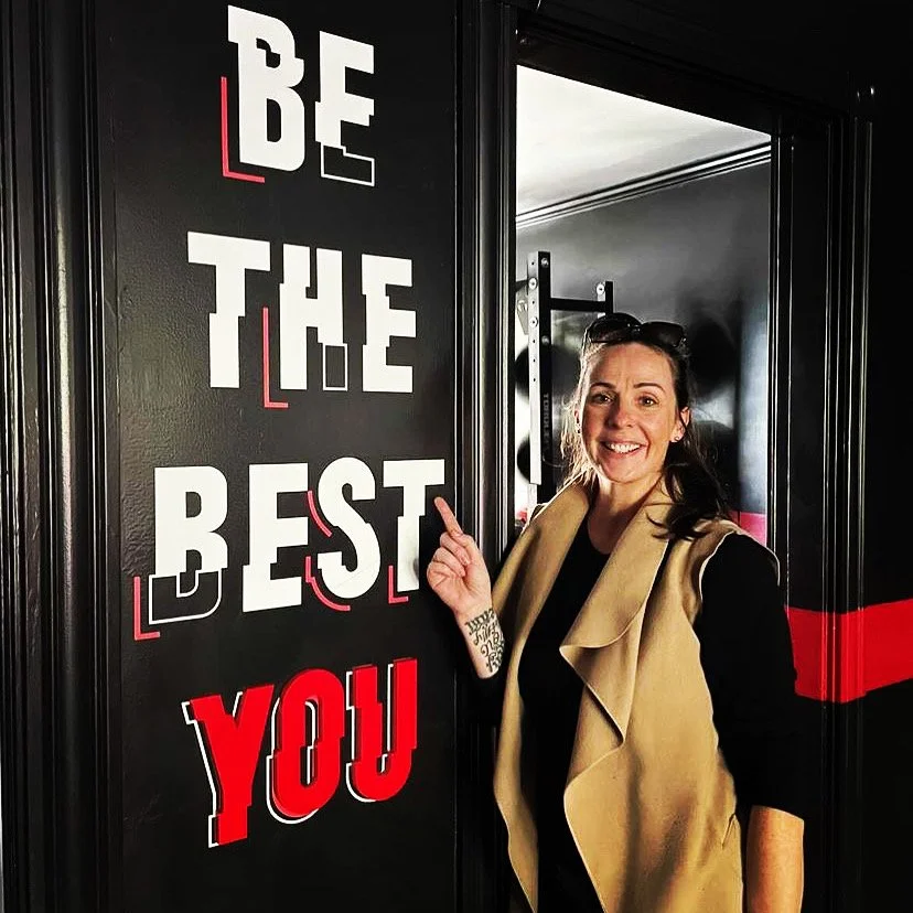 Woman smiling and pointing at a wall with the inspirational phrase "Be the Best You" in bold, white and red letters.
