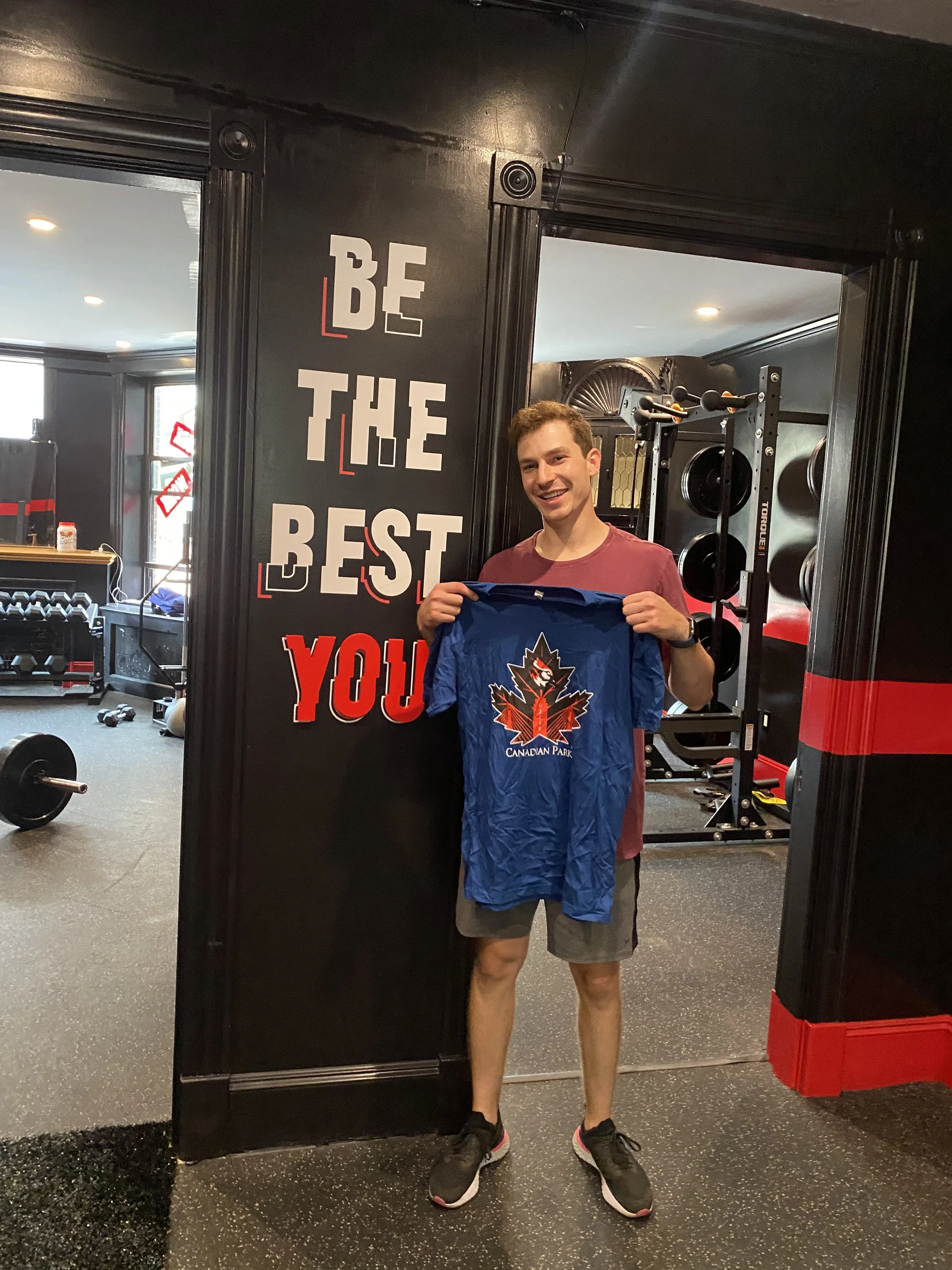 A person standing in a gym holding a blue Canadian Park T-shirt. The wall behind them has the motivational phrase "Be the Best You" in white and red letters.