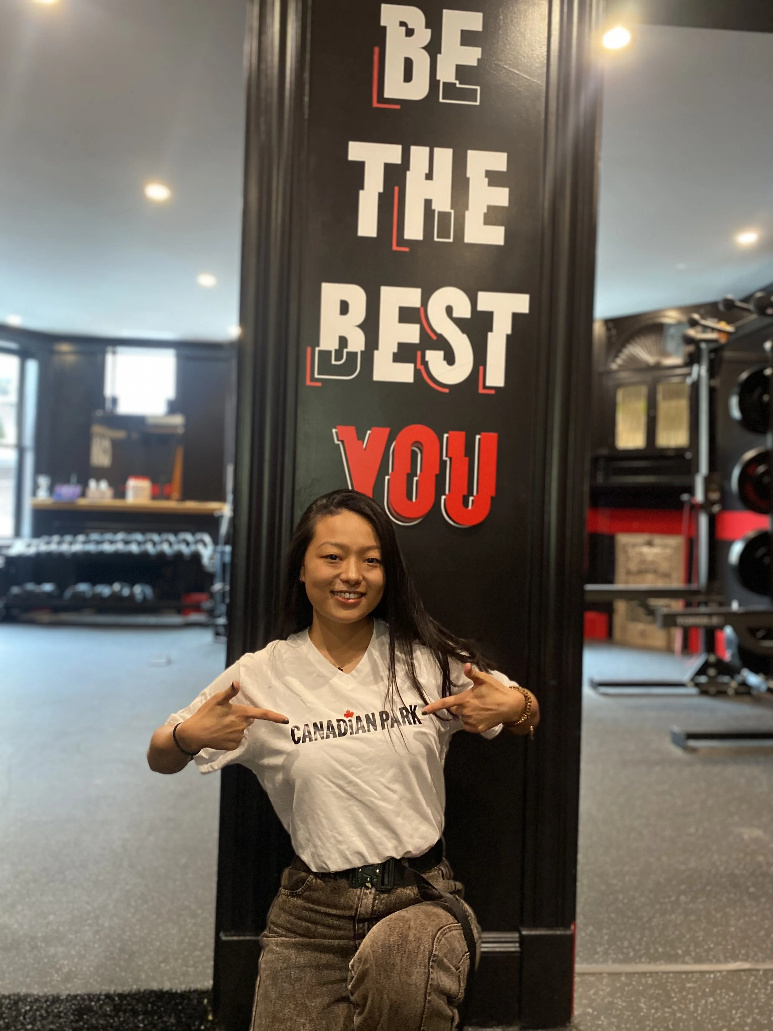 A woman kneeling in a gym, pointing at her Canadian Park t-shirt, with a sign reading 'Be The Best You' behind her.