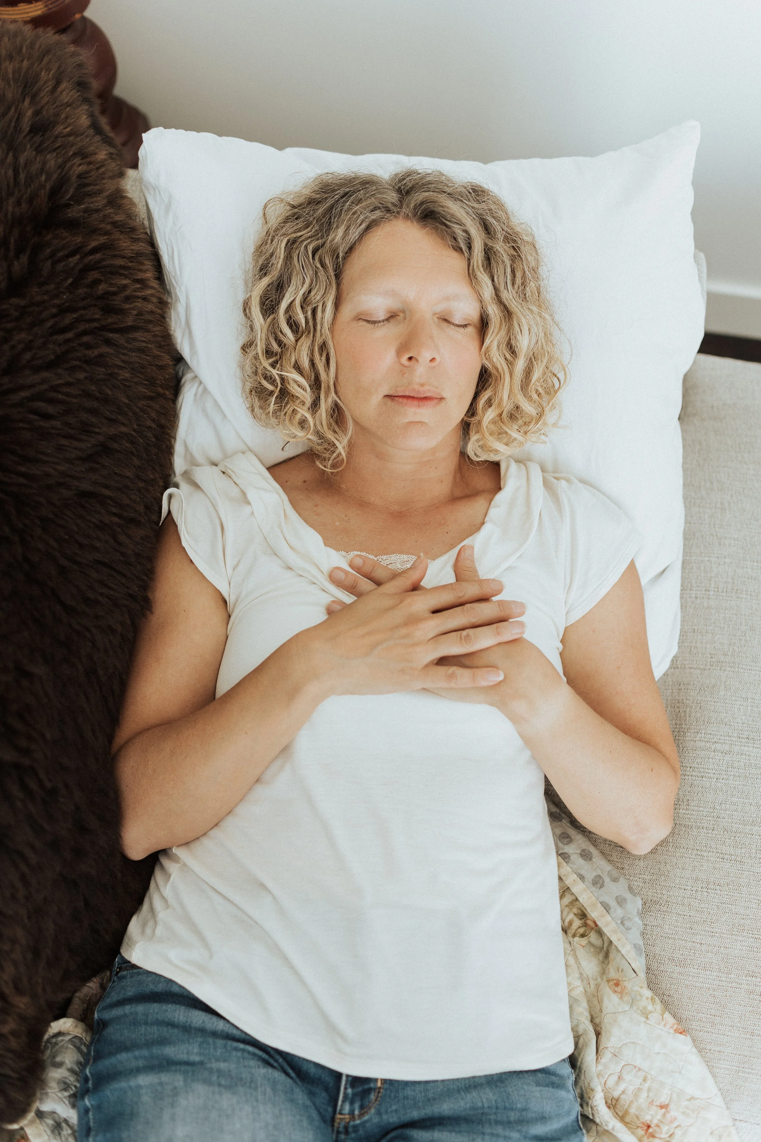 Woman lying down with hands on chest, eyes closed, wearing a white shirt and jeans, on a pillow, relaxed indoors.