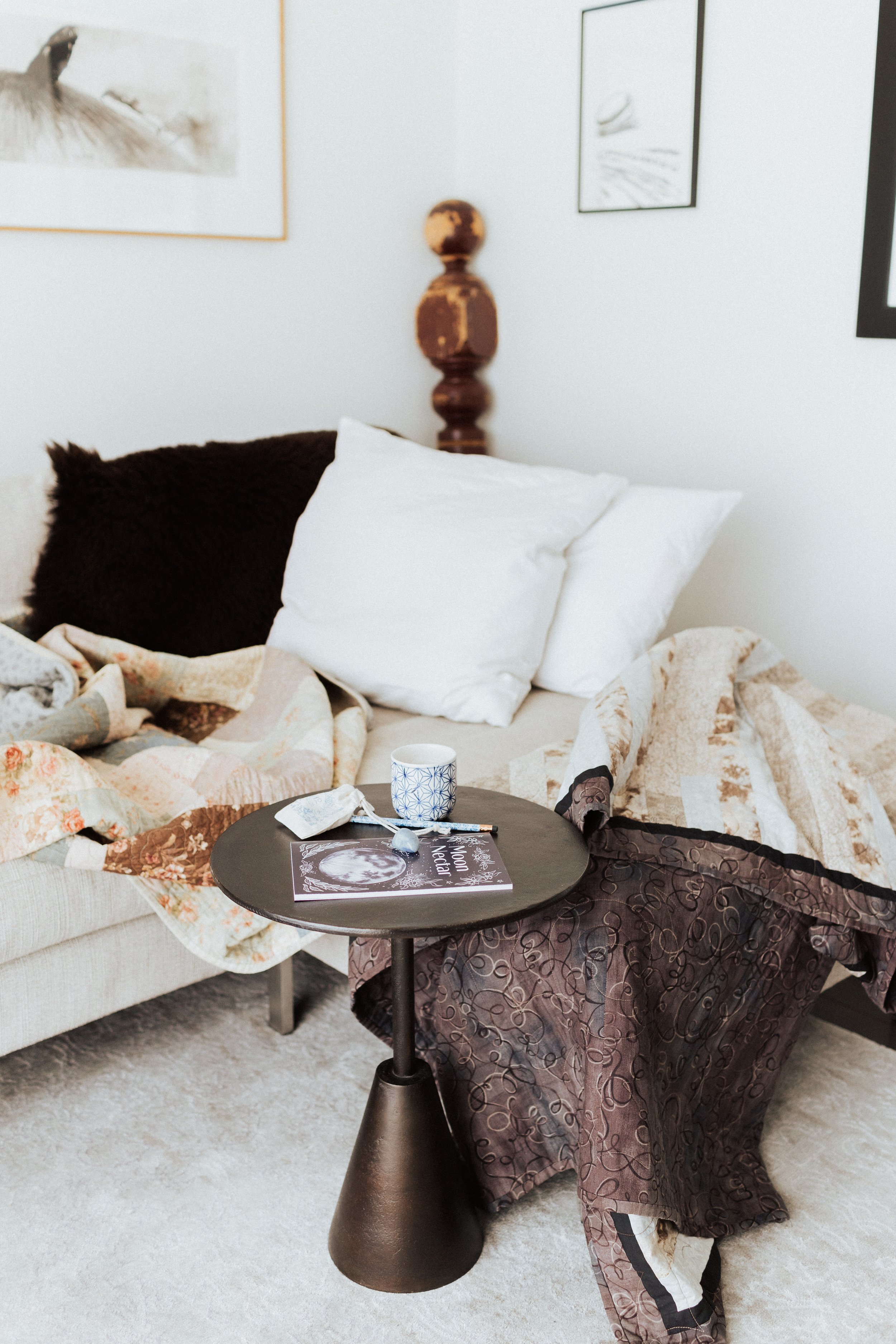 Cozy living room corner with a sofa covered in quilts and pillows, a small round table with a cup, magazine, and puzzle pieces, and framed artwork on the wall.
