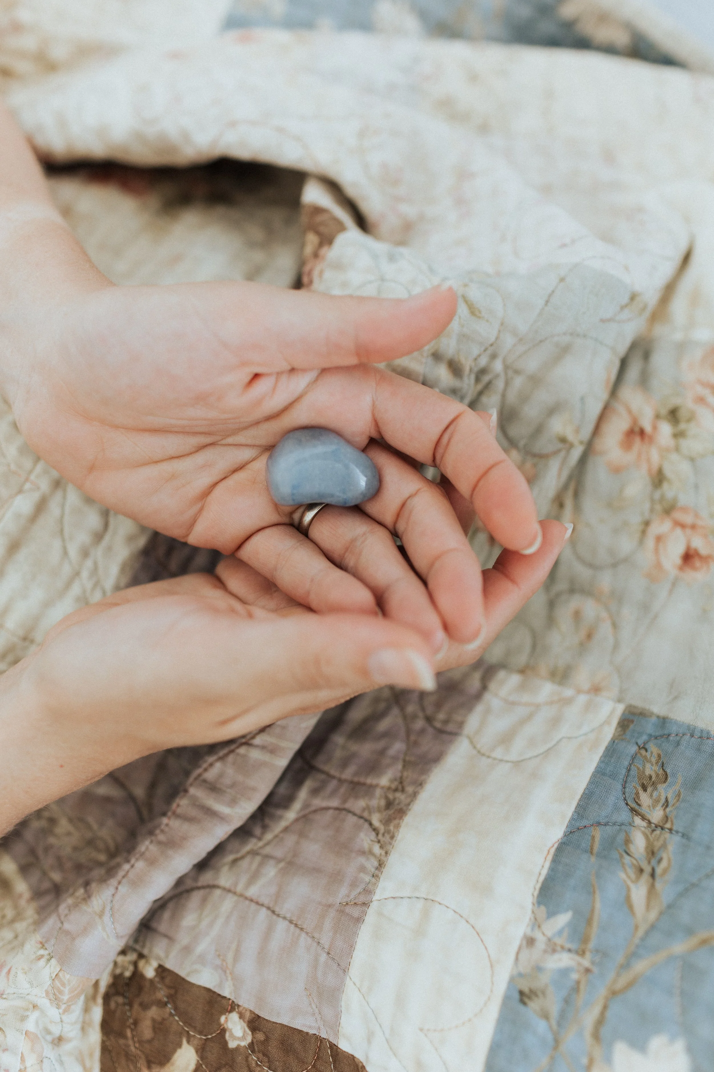 Hands holding a smooth, blue stone over a patterned quilt.