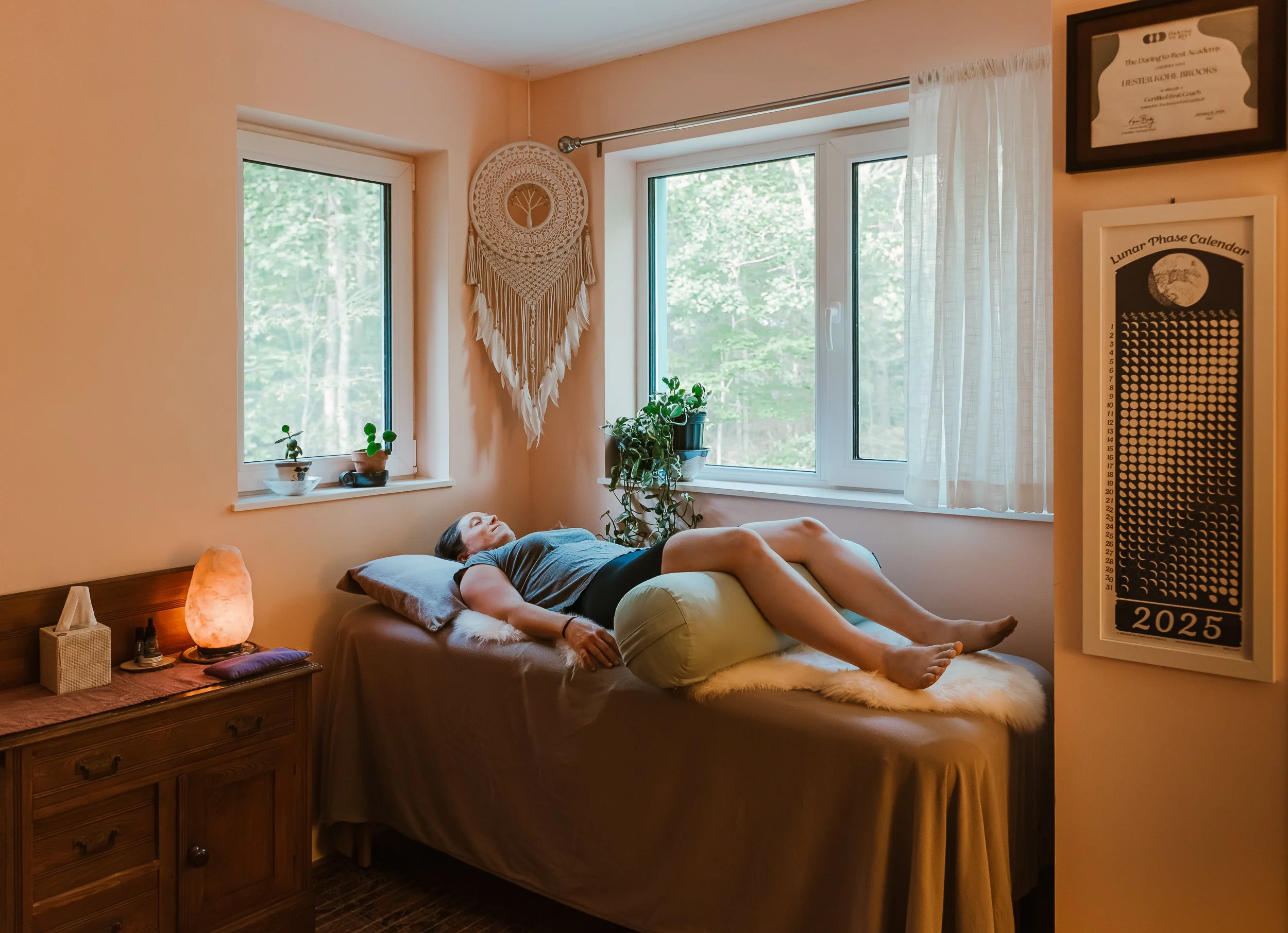 A woman lying on her side on a bed with a tan bedspread, in a cozy bedroom with natural light coming through two windows. There are houseplants on the windowsill and a dresser with a salt lamp and tissue box on it. A dreamcatcher hangs on the wall, and a lunar phase calendar is mounted on the wall beside a framed certificate.