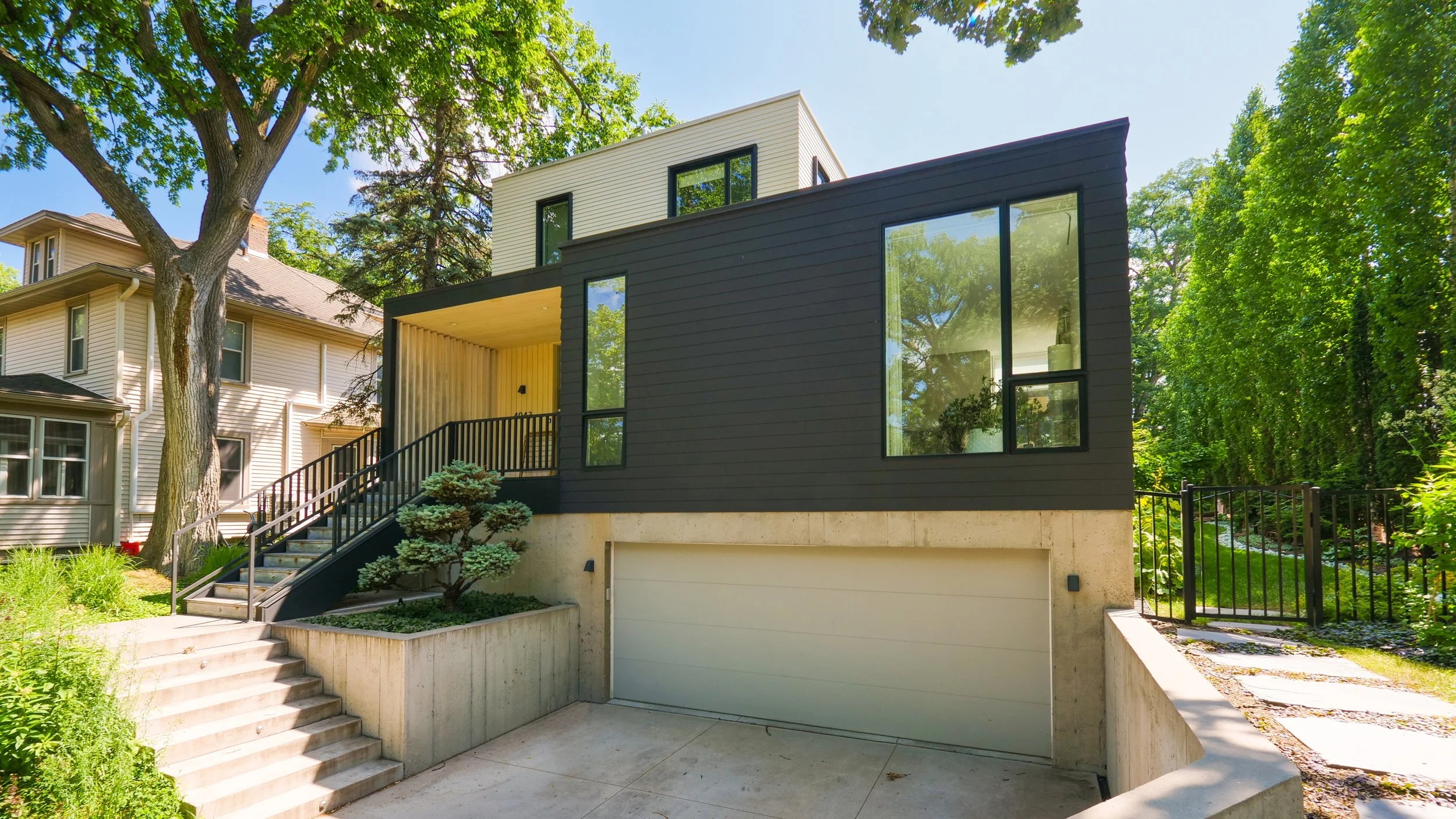 Contemporary black and white house with large glass windows, front stairs, and garage, surrounded by greenery.