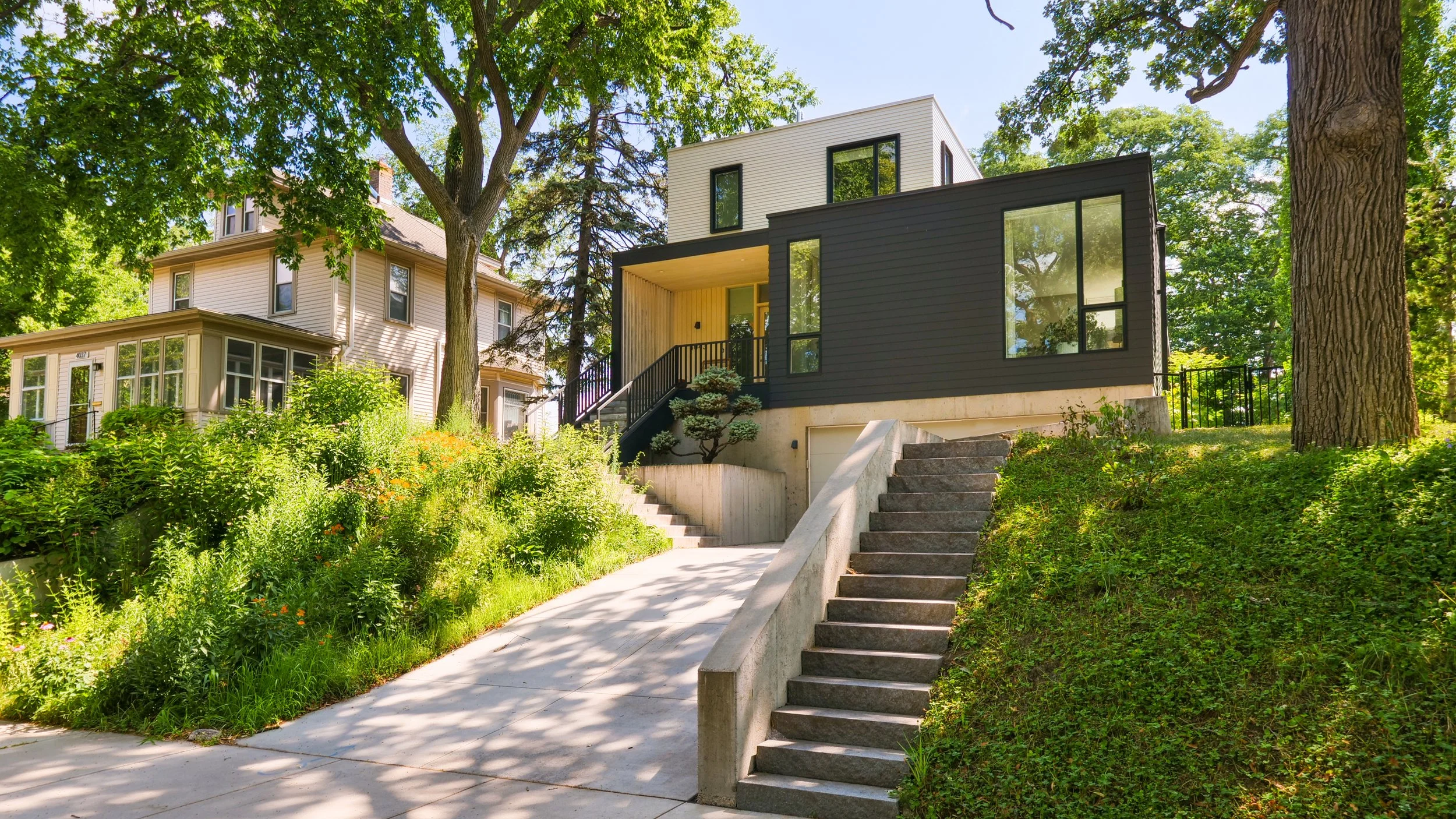 Modern black and white house with large windows, located on a lush green hillside with trees, a concrete staircase, and a driveway.