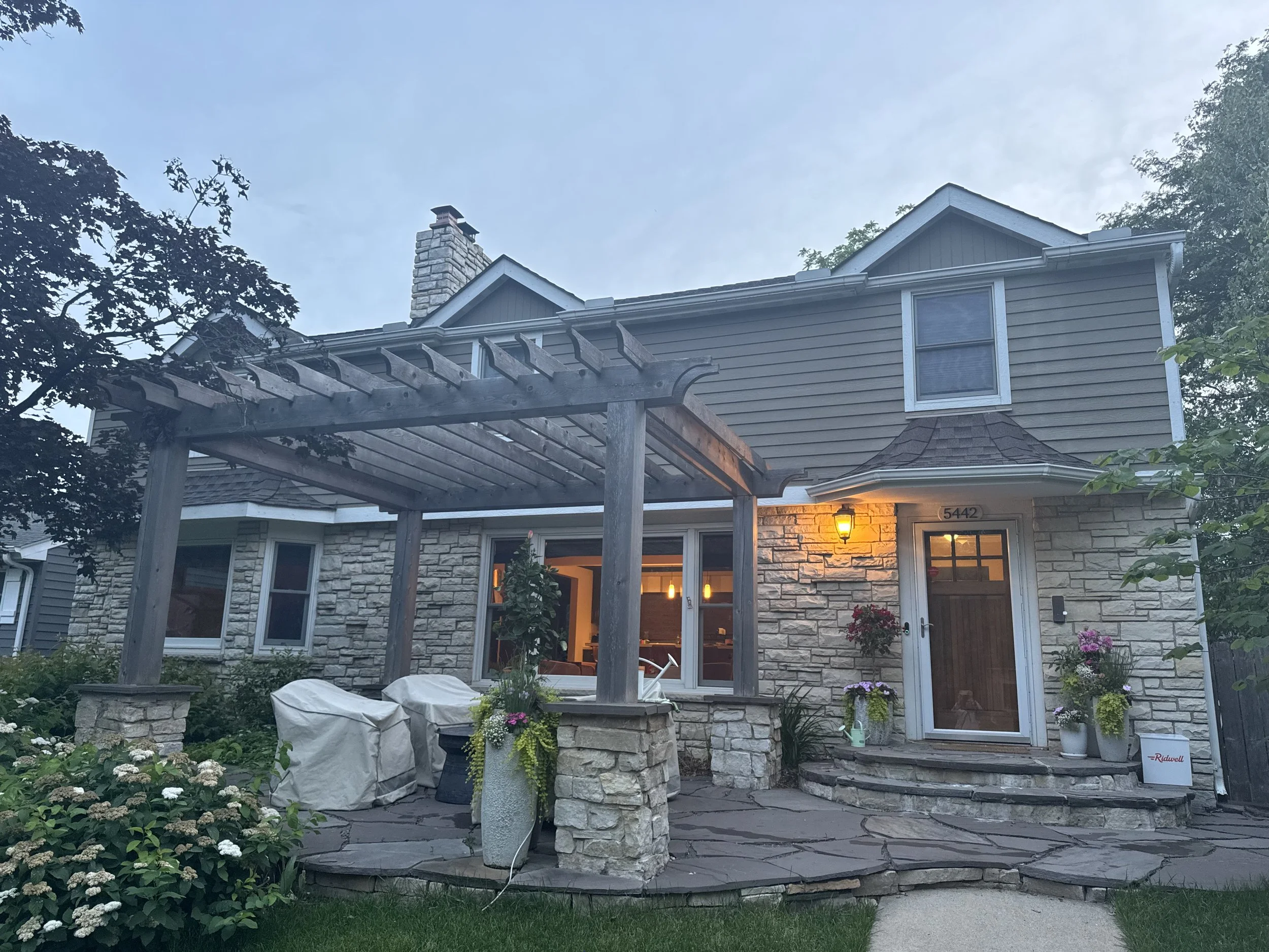 Front view of a two-story house with stone and siding exterior, a porch with stone steps, a lit porch light, and a wooden pergola over a patio with covered furniture, surrounded by plants and flowers.