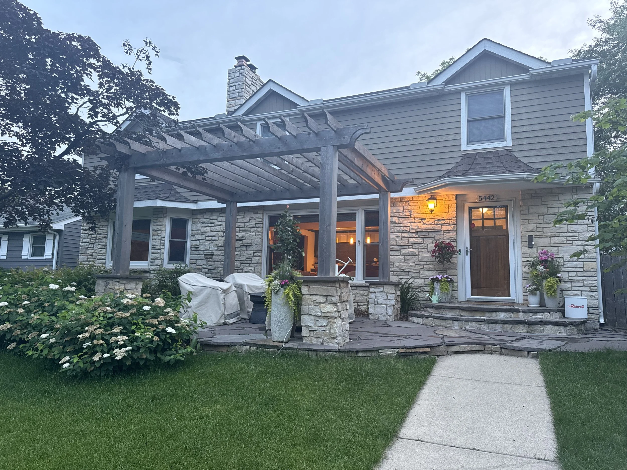 Front view of a two-story house with a stone and siding exterior, lit porch, and a circular stone walkway leading to the front door, surrounded by a well-manicured lawn and garden.