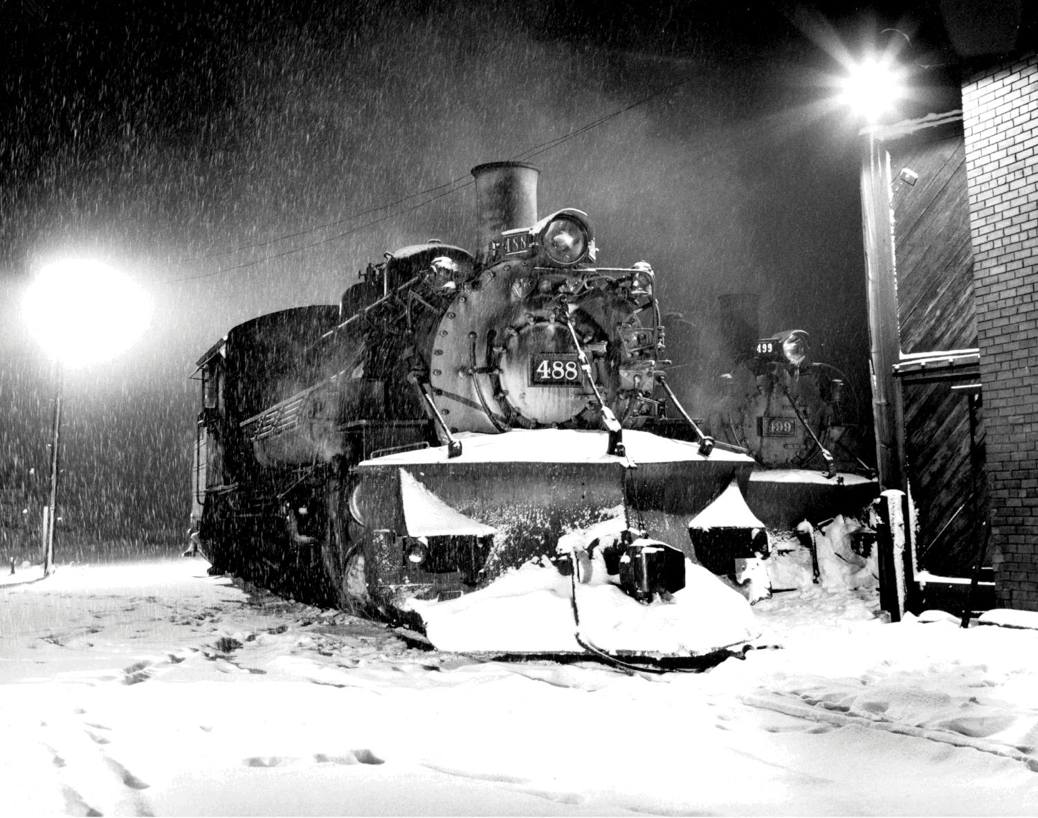 A black-and-white photograph of a steam locomotive pulling snow plow equipment, working to clear snow.