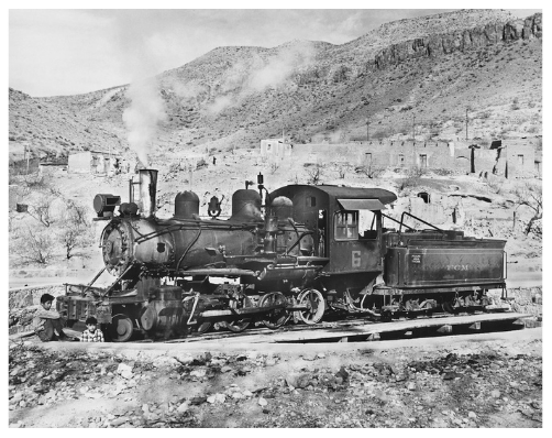 Black and white photograph of an old steam train on tracks with rocky landscape and hills in the background.