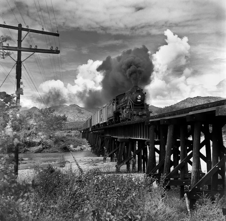A steam train traveling on a trestle bridge over a river with mountains in the background and stormy clouds in the sky.