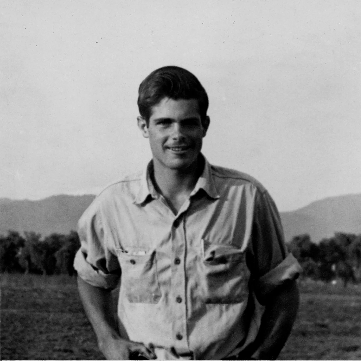 A young Frank Barry  with dark hair standing outdoors in a field with mountains in the background, smiling at the camera, in black and white.