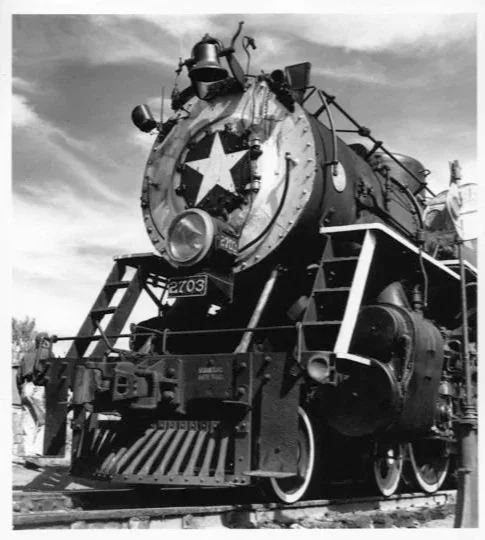 Black and white photograph of a vintage steam locomotive train, with a large star emblem on the front, moving on railroad tracks during daytime.