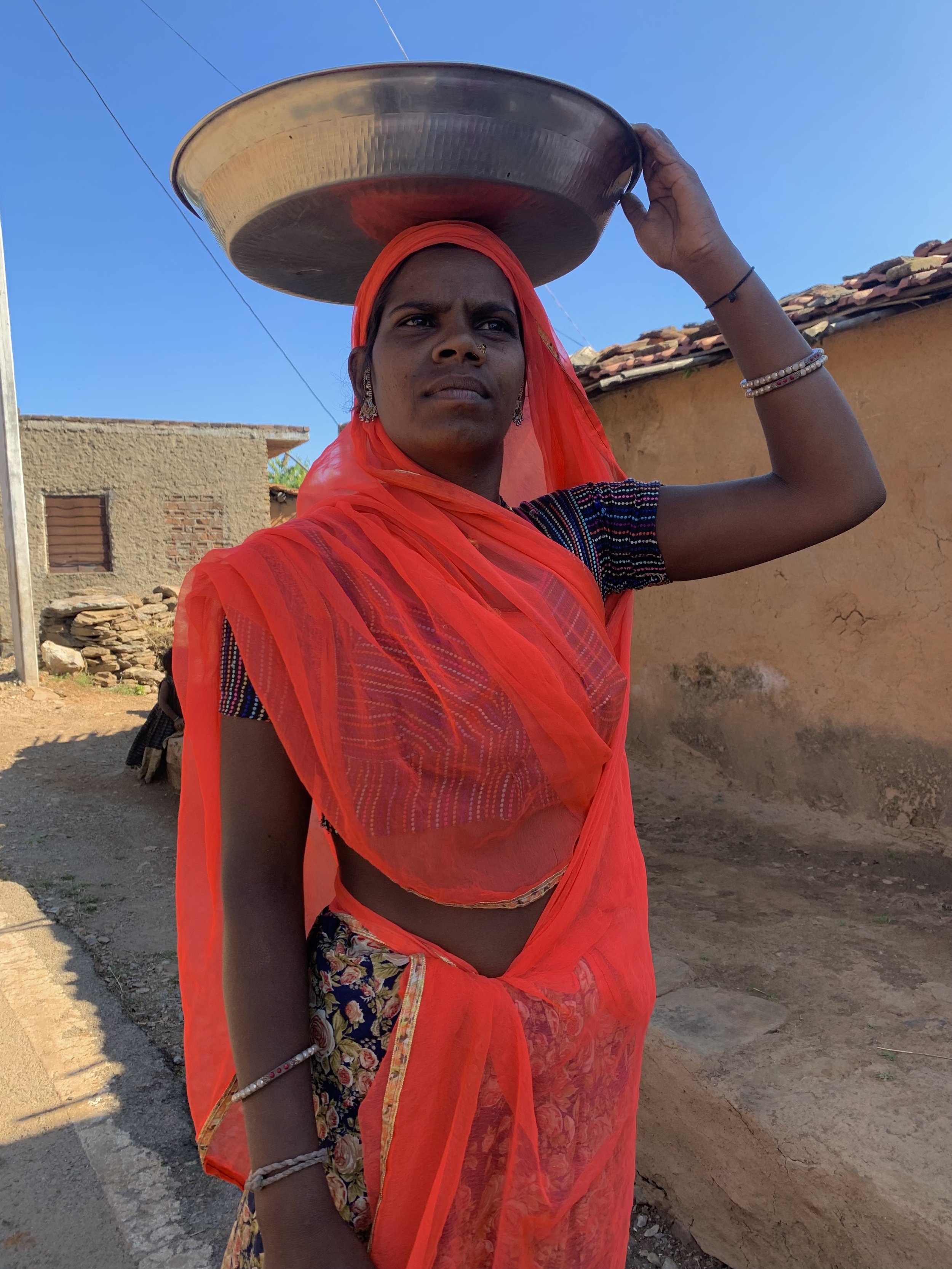 A woman in traditional Indian attire, wearing a bright orange saree and headscarf, balances a large metal tray on her head while walking outdoors on a sunny day.