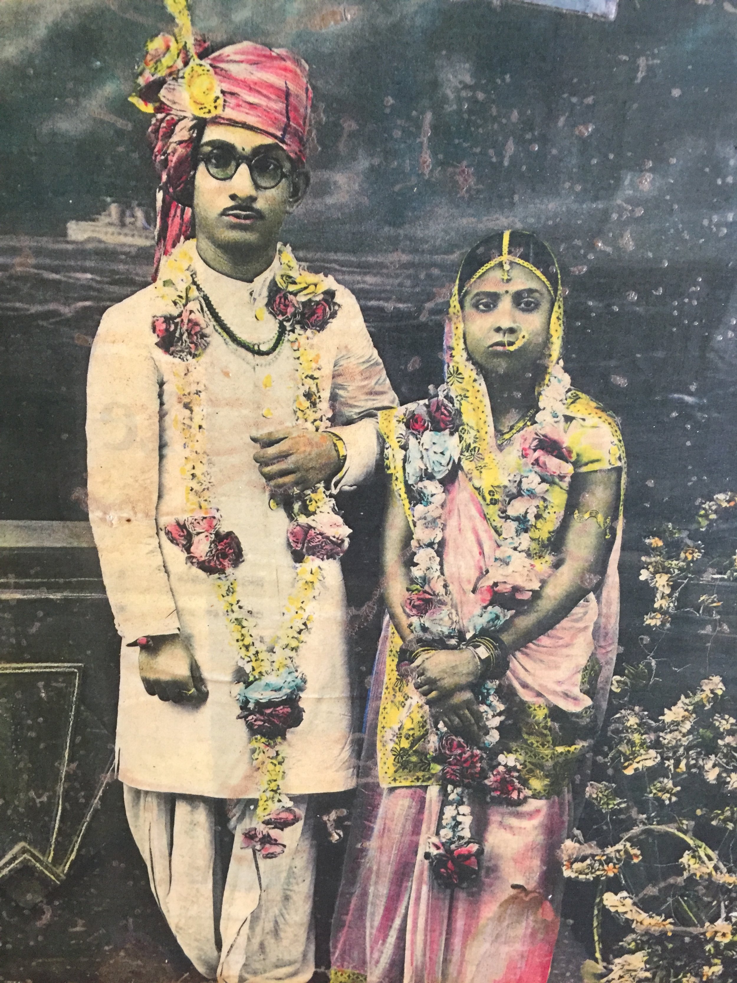 A vintage photo of a man and woman dressed in traditional Indian wedding attire, standing outdoors with a dark cloudy sky in the background, both adorned in flower garlands.