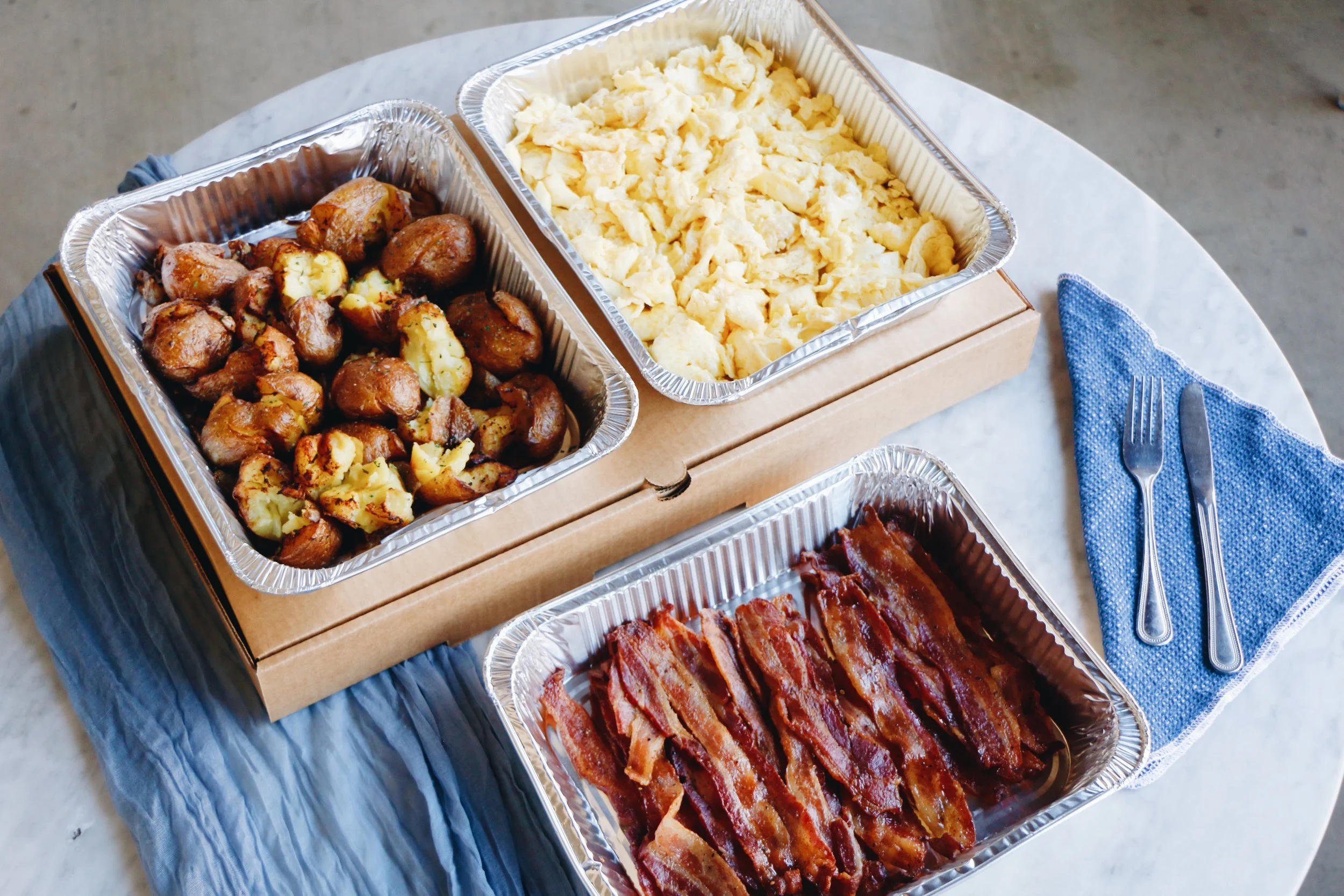 Catering breakfast spread from Prep & Pastry featuring aluminum trays of roasted breakfast potatoes, scrambled eggs, and crispy bacon next to a blue linen napkin and silverware..jpg